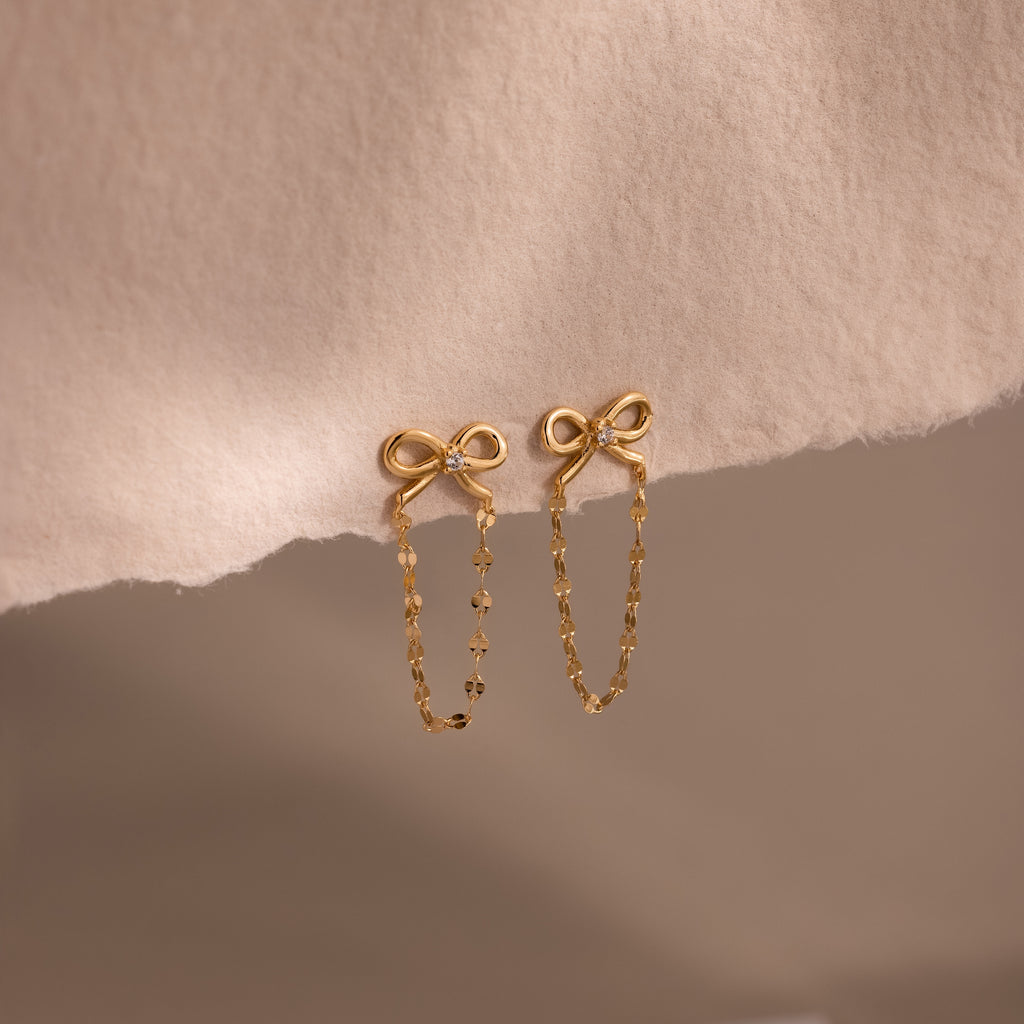 Bow Chain Studs featuring delicate chain dangles, shown on a soft beige fabric—ideal for fans of feminine and vintage-inspired jewelry styles.