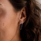 A womans ear wears Bow Chain Studs in Sterling Silver, featuring a delicate silver bow with a vintage-inspired design and a dangling chain.