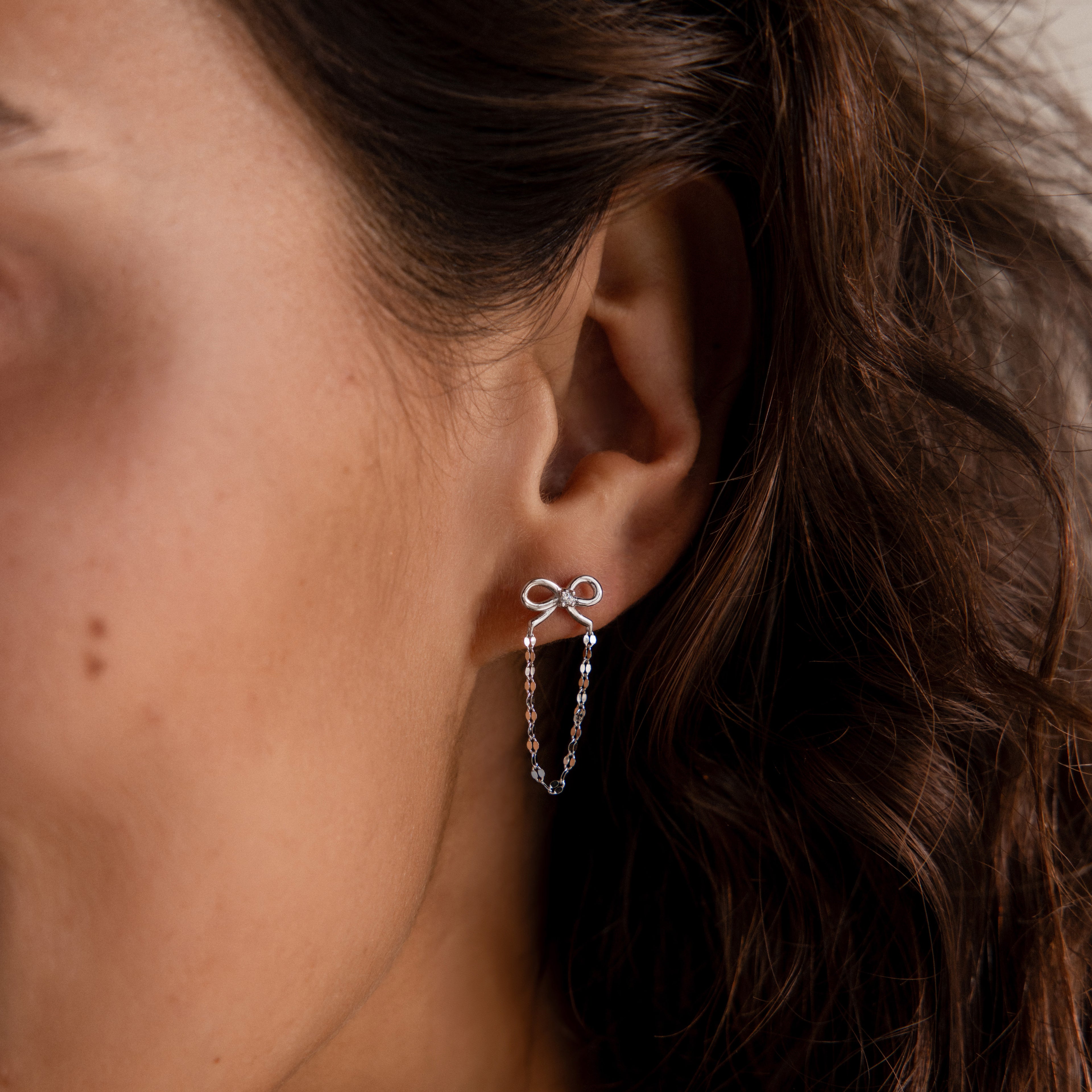 A womans ear wears Bow Chain Studs in Sterling Silver, featuring a delicate silver bow with a vintage-inspired design and a dangling chain.