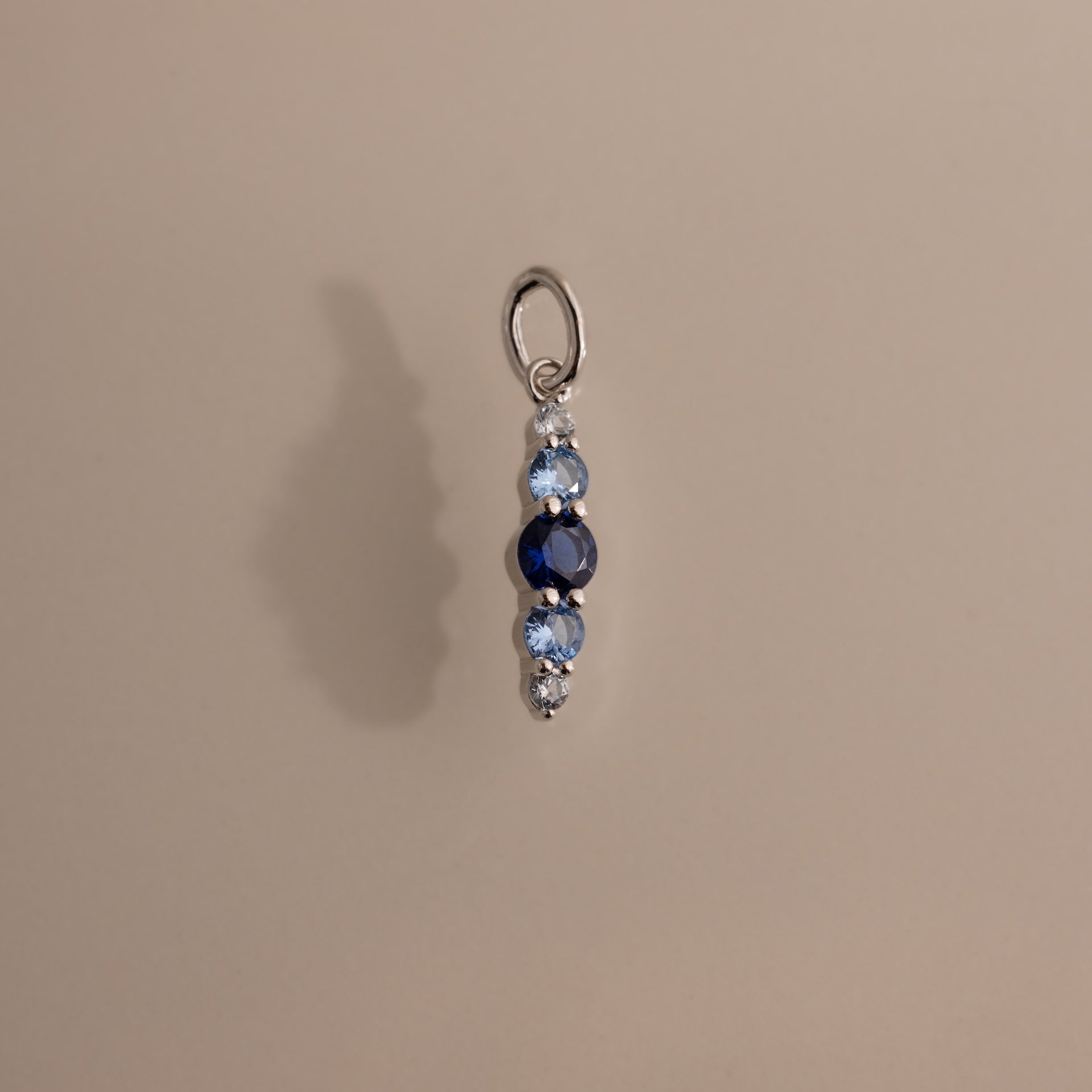 The Ombre Lace Blue Zircon Charm is a silver pendant featuring three blue gemstones in varying shades, arranged vertically against a beige background.