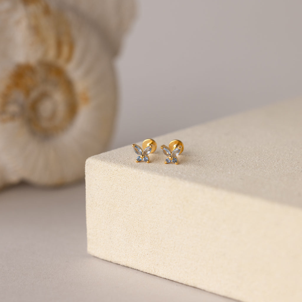Butterfly Blue Zircon Flatback Studs, adorned with small gemstones and a flat back stud design, are elegantly displayed on a beige textured surface.