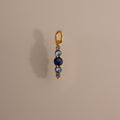 The Ombre Lace Blue Zircon Charm, a gold pendant with a vertical row of blue and silver beads, is photographed on a beige background.