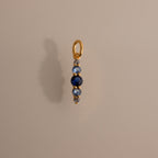 The Ombre Lace Blue Zircon Charm, a gold pendant with a vertical row of blue and silver beads, is photographed on a beige background.