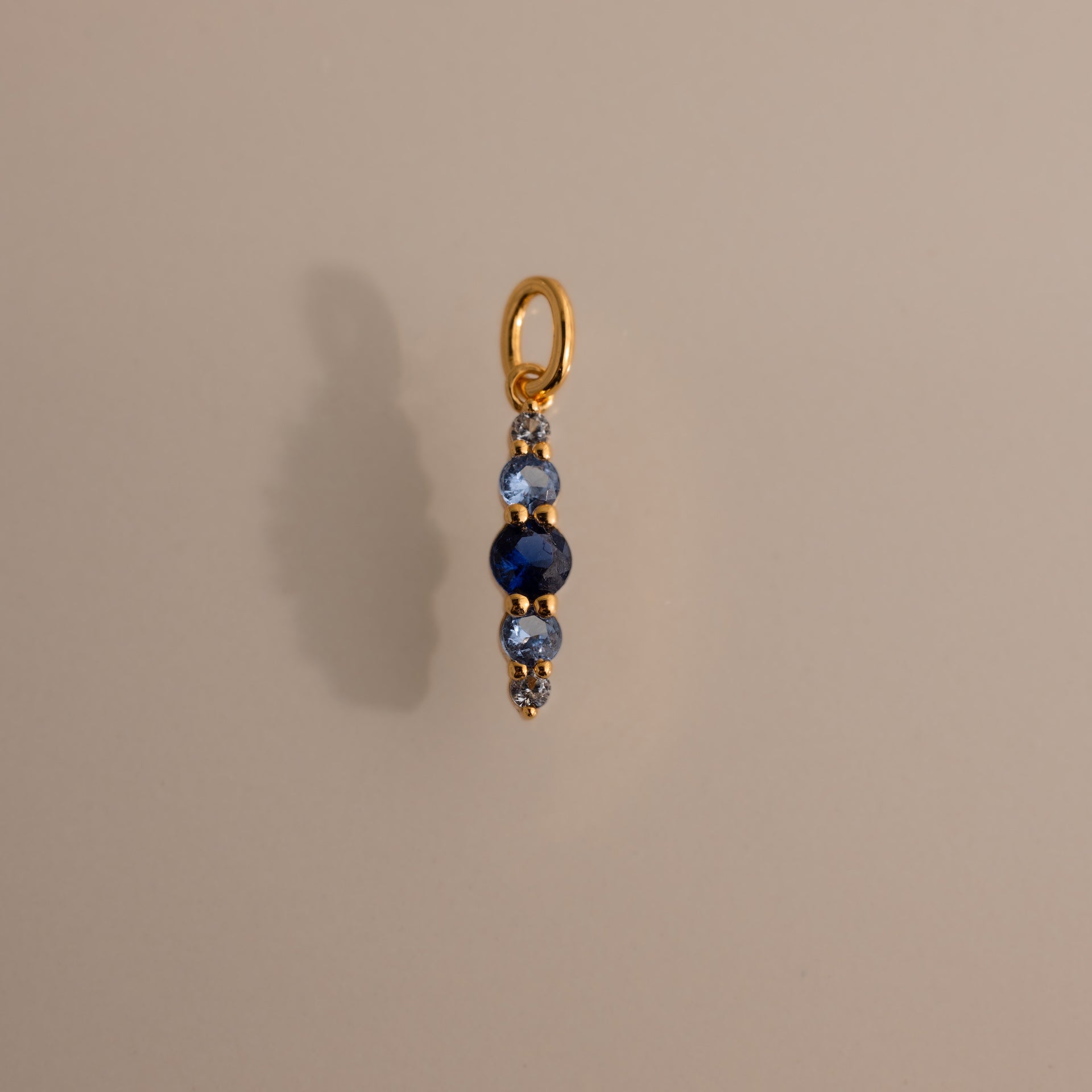 The Ombre Lace Blue Zircon Charm, a gold pendant with a vertical row of blue and silver beads, is photographed on a beige background.