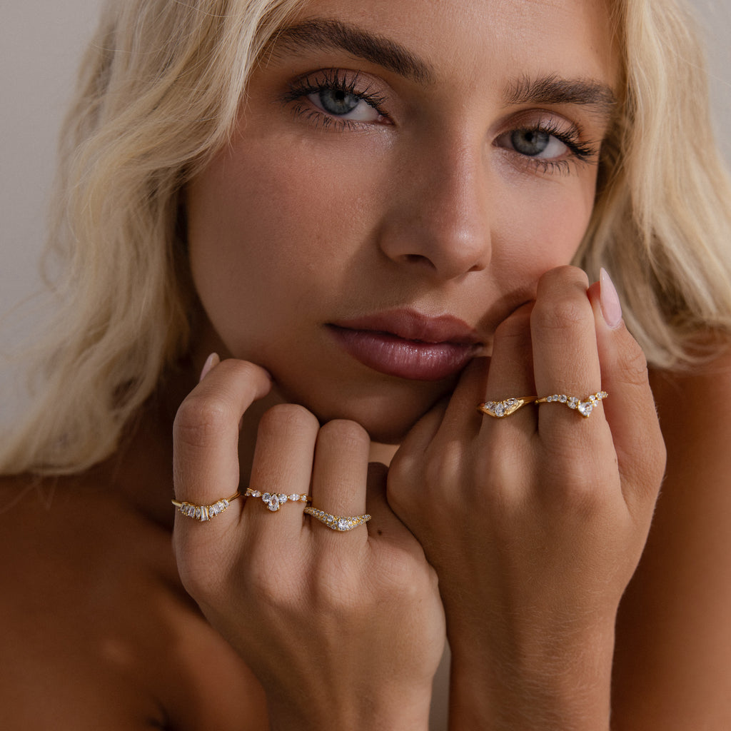 Model resting her face on her hands, wearing multiple gold diamond rings, with the gold pavé diamond curve ring highlighted on her left hand, featuring a delicate arc lined with sparkling pavé-set diamonds.