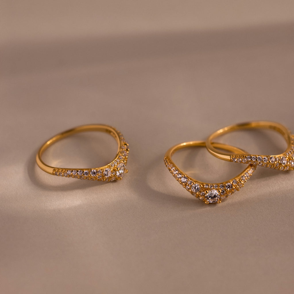 Gold pavé diamond curve rings displayed on a neutral surface, featuring delicate curved bands set with shimmering pavé diamonds and a central round diamond.