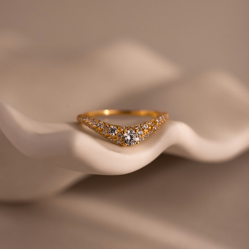 Gold pave curve ring with a round brilliant cut center diamond, accented by shimmering pave set stones along the band, displayed on a sculpted neutral background.