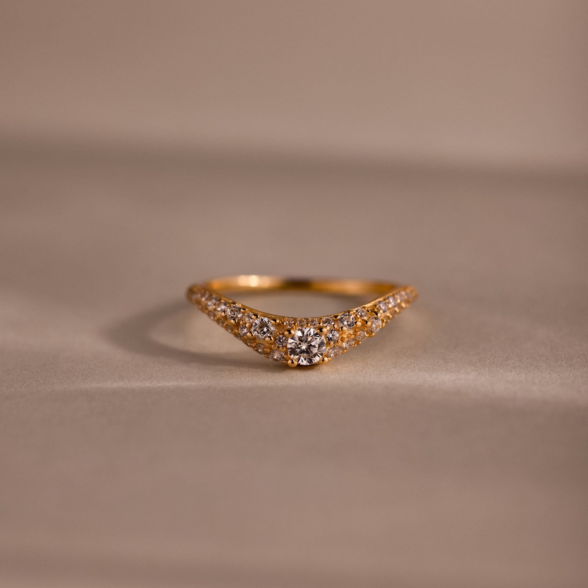 Gold pave curve ring with a round center diamond and shimmering pave set stones along the band, displayed on a soft neutral background.
