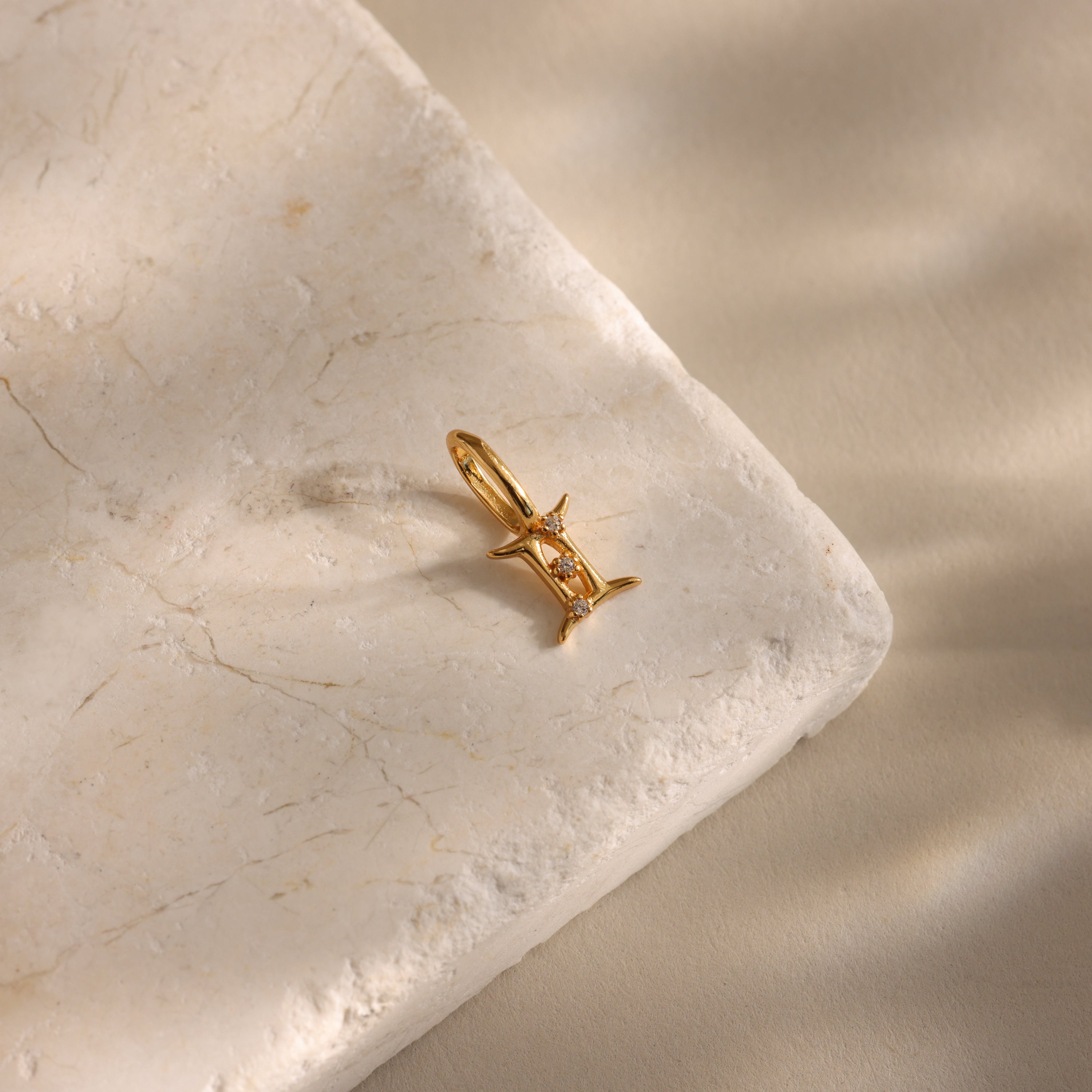 The Zodiac Charm, a gold pendant with a small gemstone, sits on a textured light stone surface with soft shadows, showcasing the elegance of celestial jewelry.