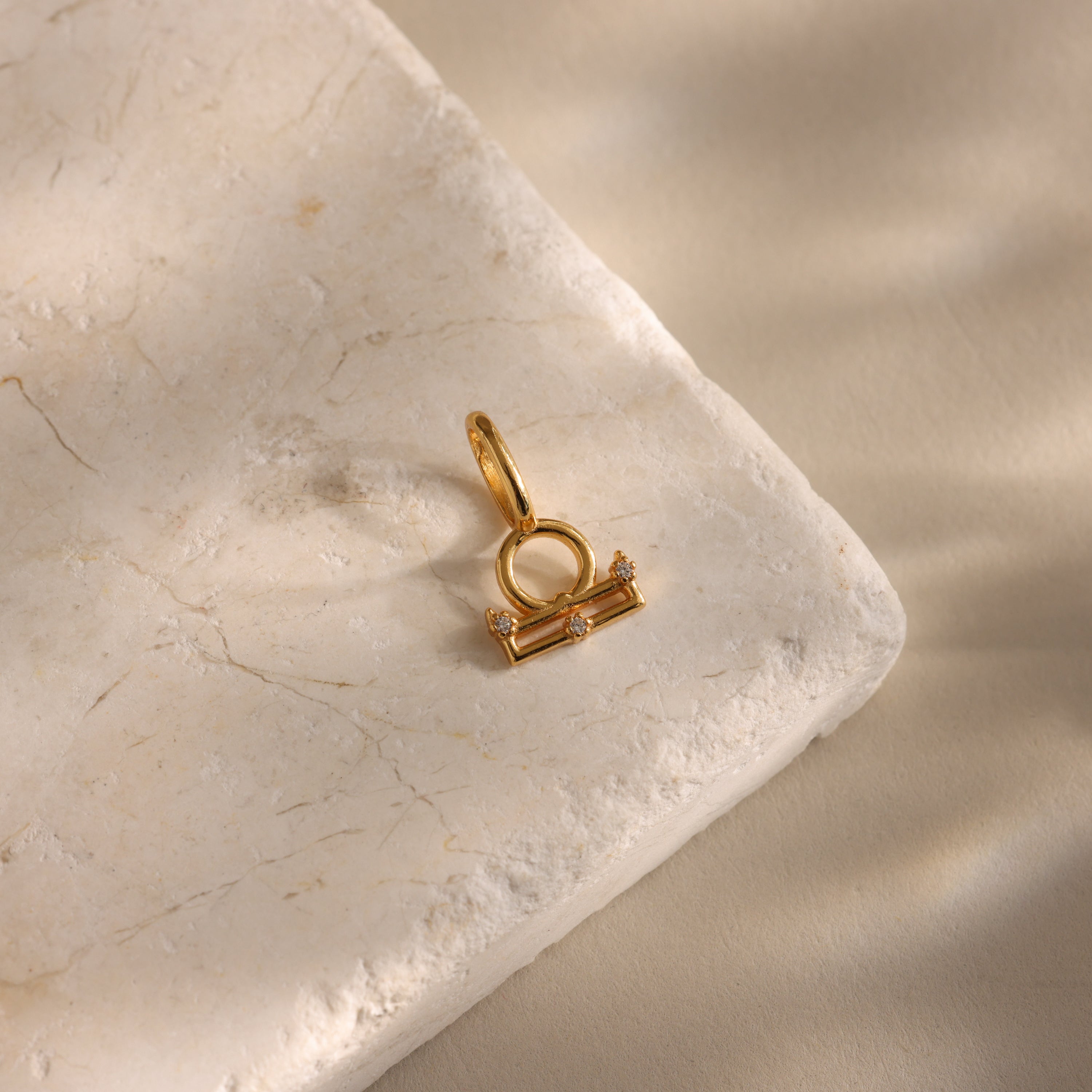 The Zodiac Charm pendant, adorned with small gems, rests on a marble slab in soft natural light—a stunning piece of celestial jewelry.
