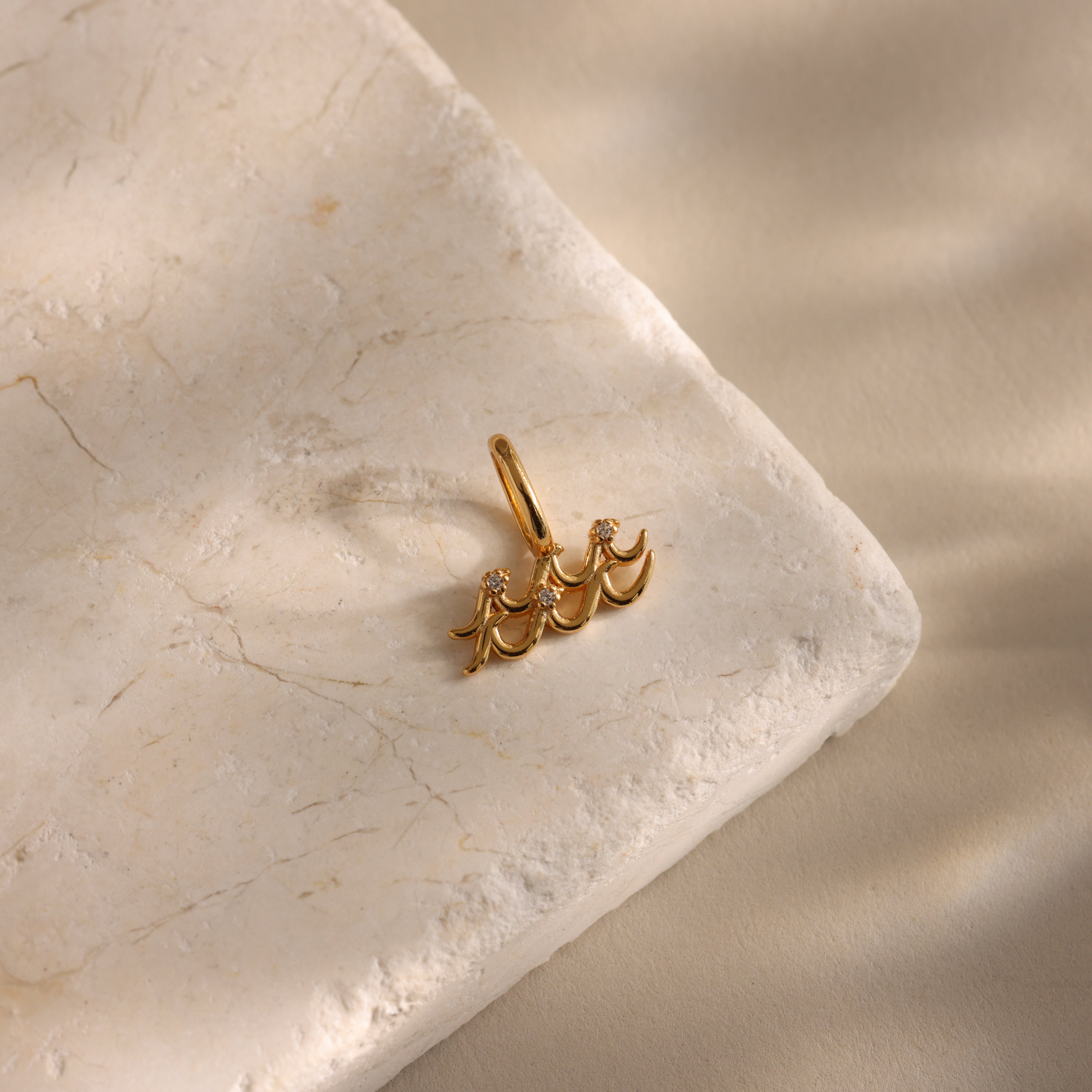 The Zodiac Charm gold earring features three crescent shapes and small diamonds, creating a celestial glow as it rests elegantly atop a textured beige stone surface.