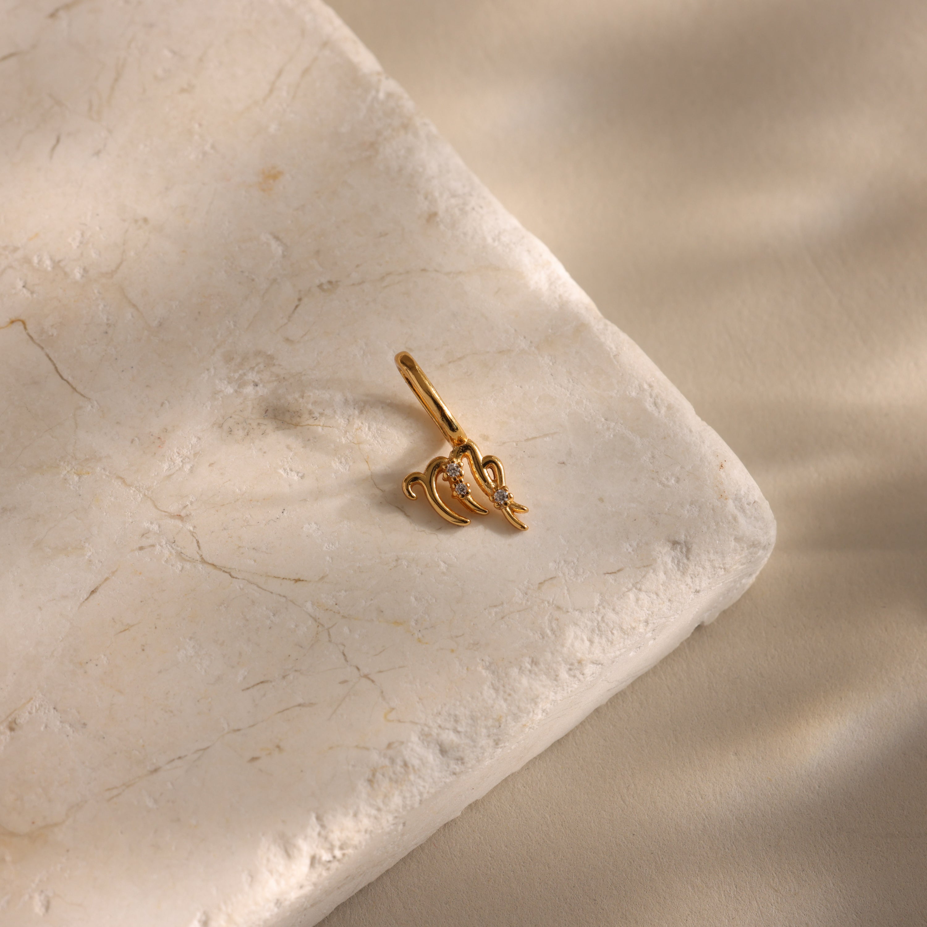 The Zodiac Charm, a small gold and diamond ear cuff inspired by celestial jewelry, rests on a textured light stone surface with soft shadows.
