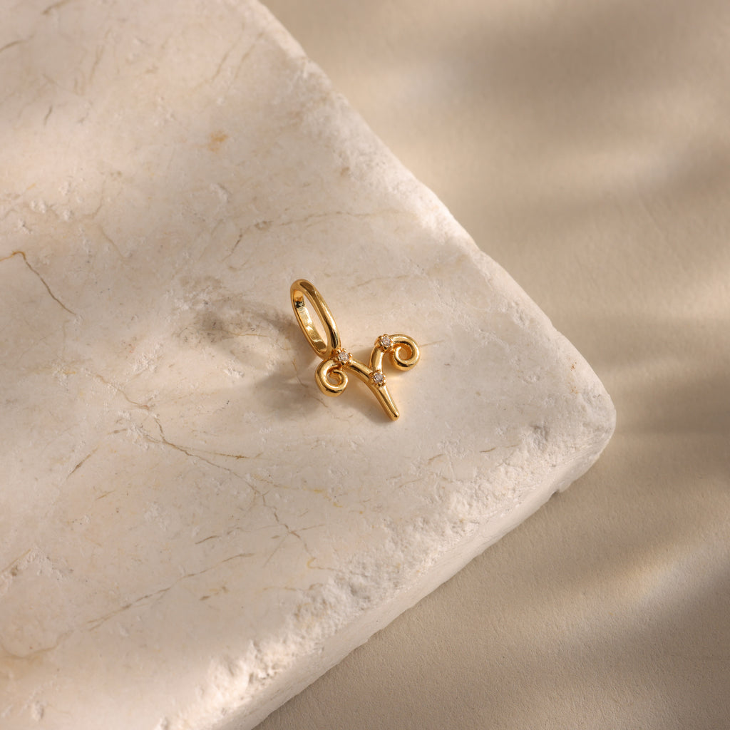 The Zodiac Charm, a gold Aries pendant, rests on a textured beige stone with soft shadows, adding celestial elegance to your jewelry collection.