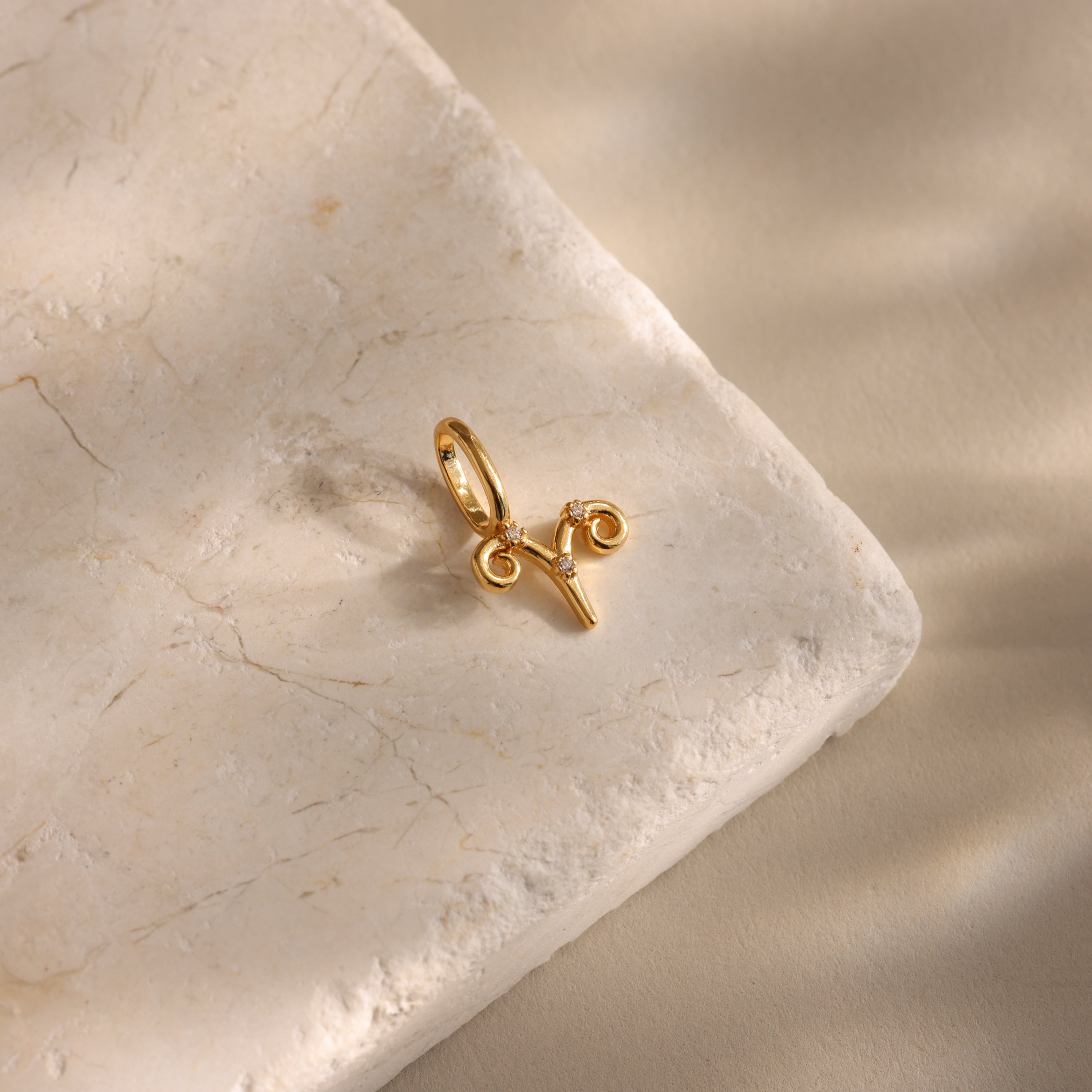 The Zodiac Charm, a gold Aries pendant, rests on a textured beige stone with soft shadows, adding celestial elegance to your jewelry collection.