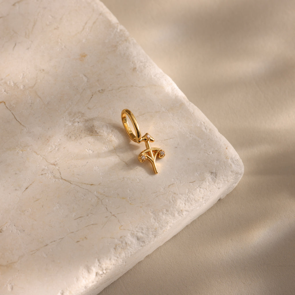 The Zodiac Charm is a gold pendant featuring a cross design with small stones, inspired by celestial jewelry, and shown on a textured beige stone surface.