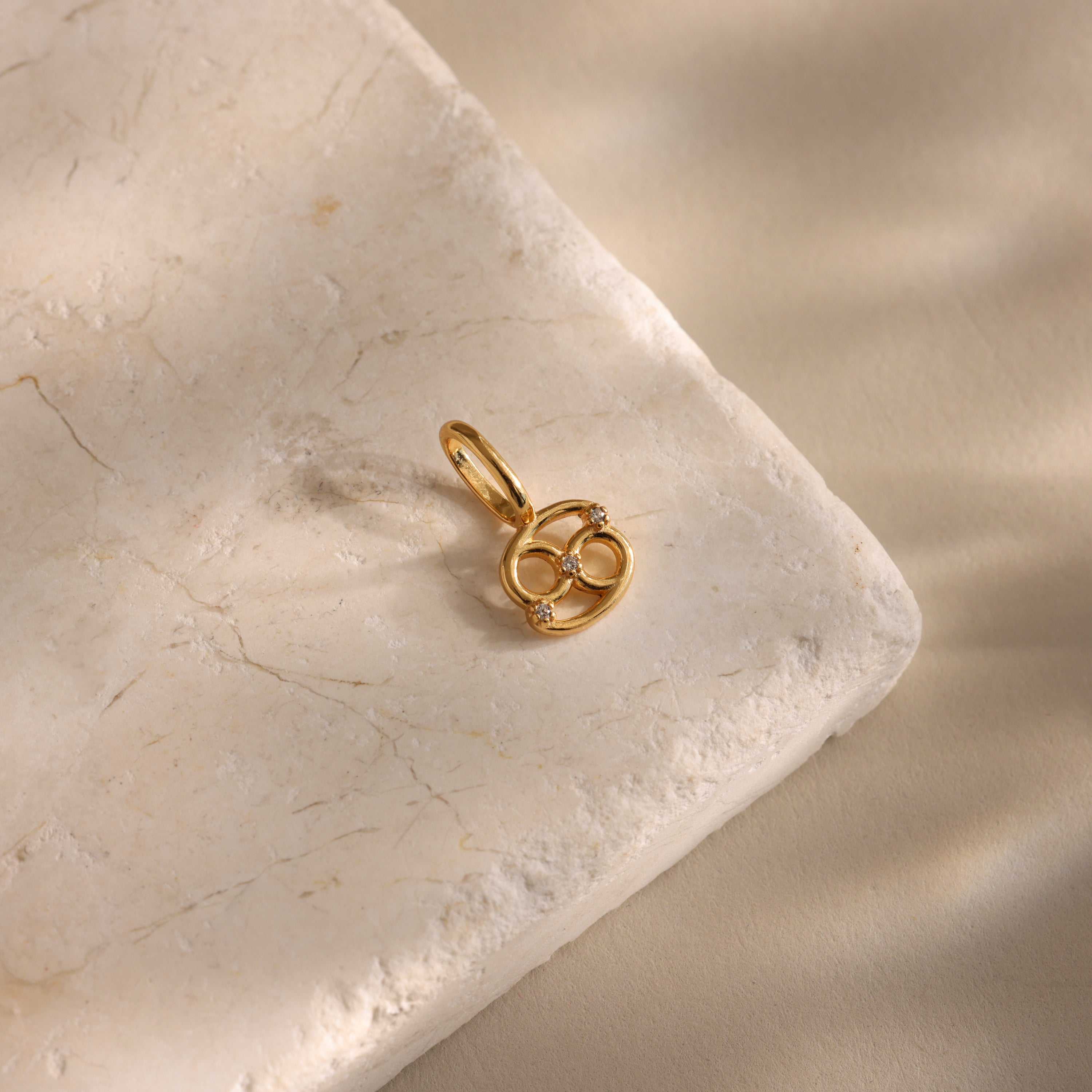 The Zodiac Charm, an infinity-shaped gold pendant with small diamonds, glimmers elegantly on a light textured stone surface.