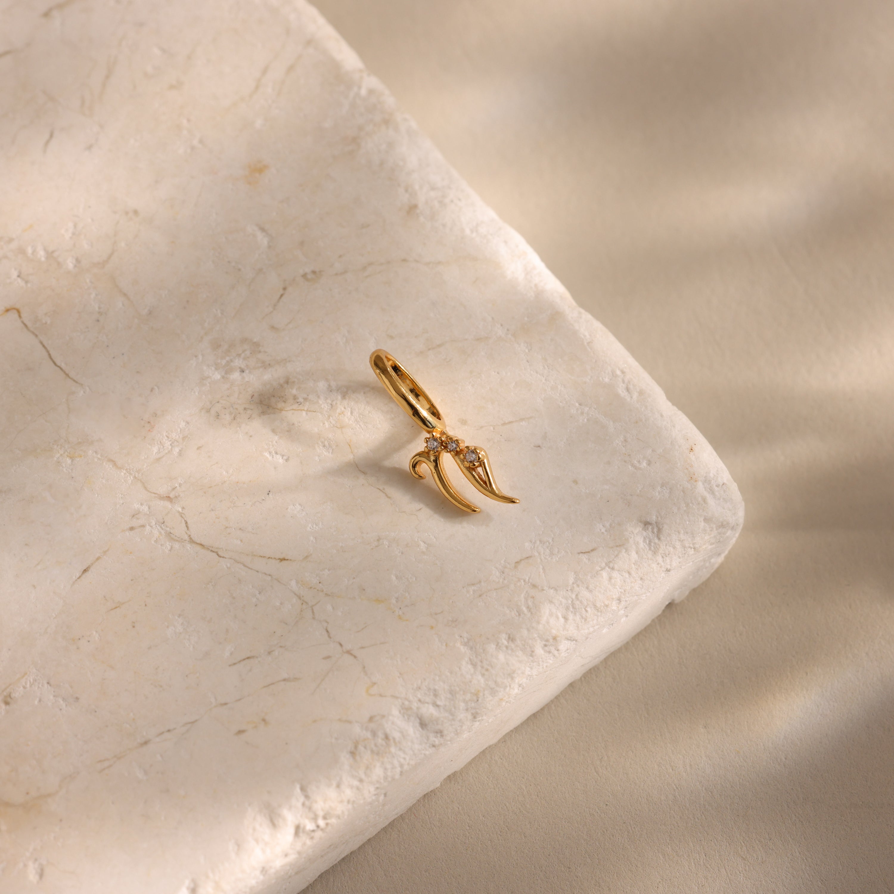 The Zodiac Charm, a gold claw-shaped pendant, is displayed on a textured, light-colored stone surface under soft lighting.