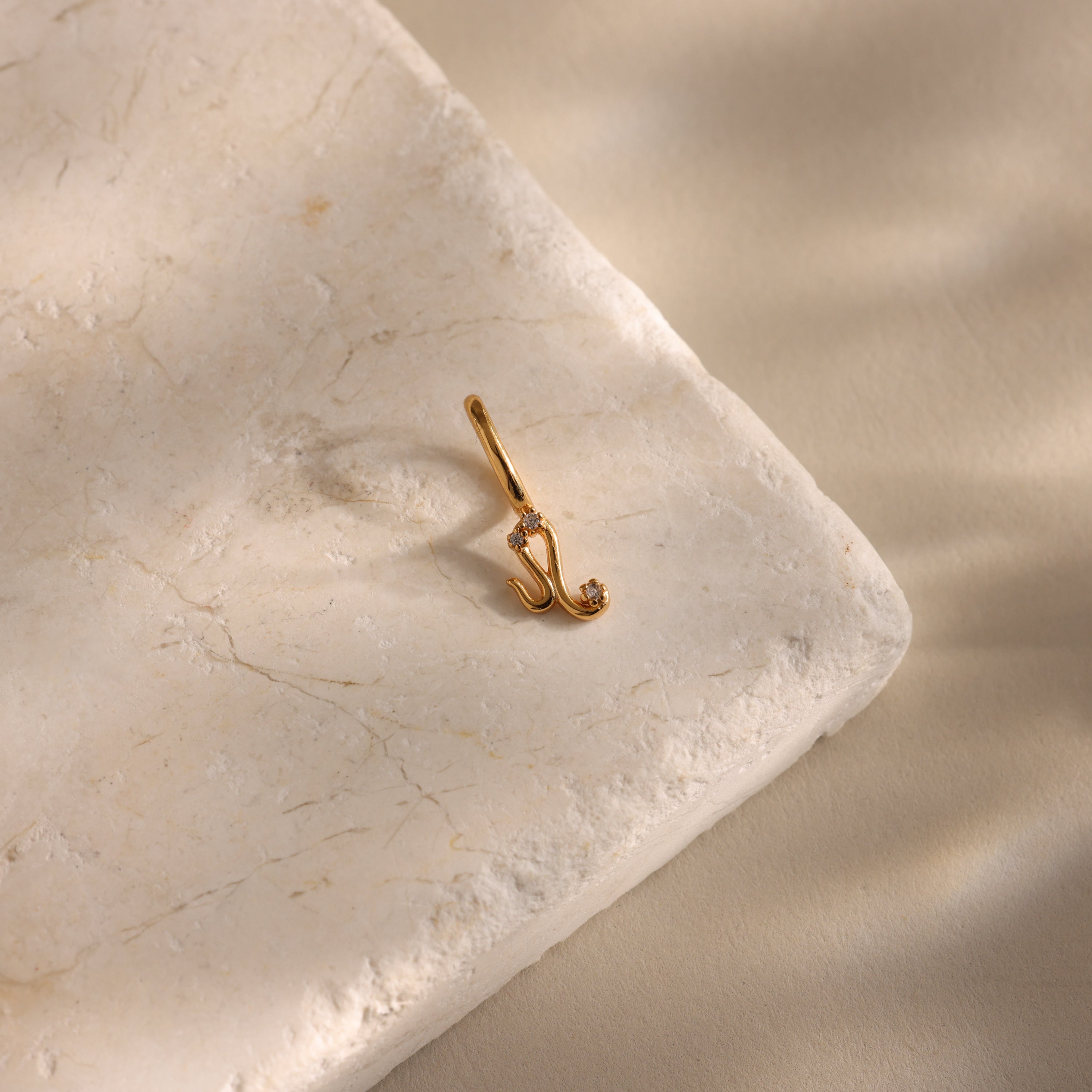 Zodiac Charm: Gold pendant shaped like the letter J, accented with a small gemstone and displayed on a textured stone surface—ideal for personalized celestial jewelry collections.