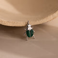 The Malachite Beetle Charm in silver lying upright, showing its vivid green wing casings and polished gold legs and crown.