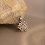 A close-up image of a silver Opal Sun Charm resting against a textured beige stone. The charm features a radiant, sculpted sunburst design with an iridescent opal stone at its center, glowing with hues of orange, green, and blue. The charm has a small polished gold bail for attachment.