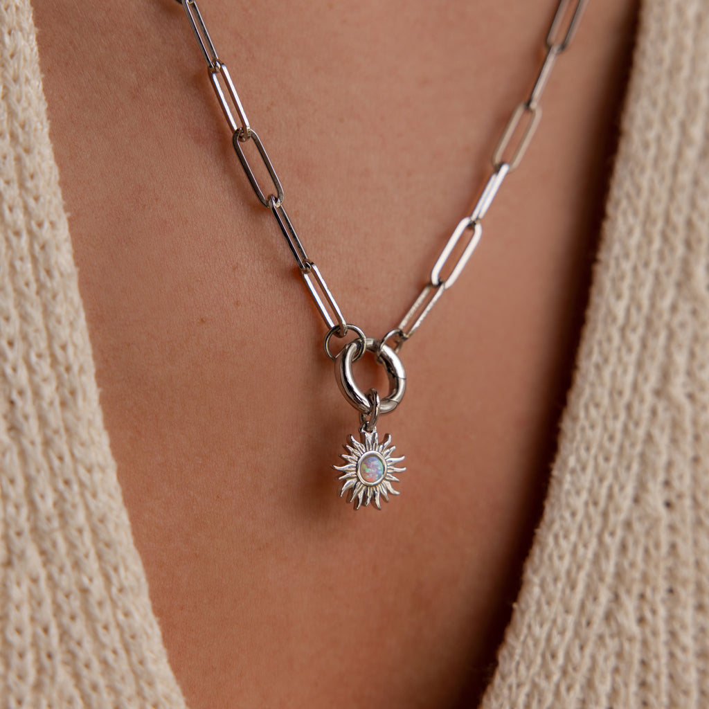 Silver sun charm with an opal center on a paperclip chain necklace, worn over a textured beige top.