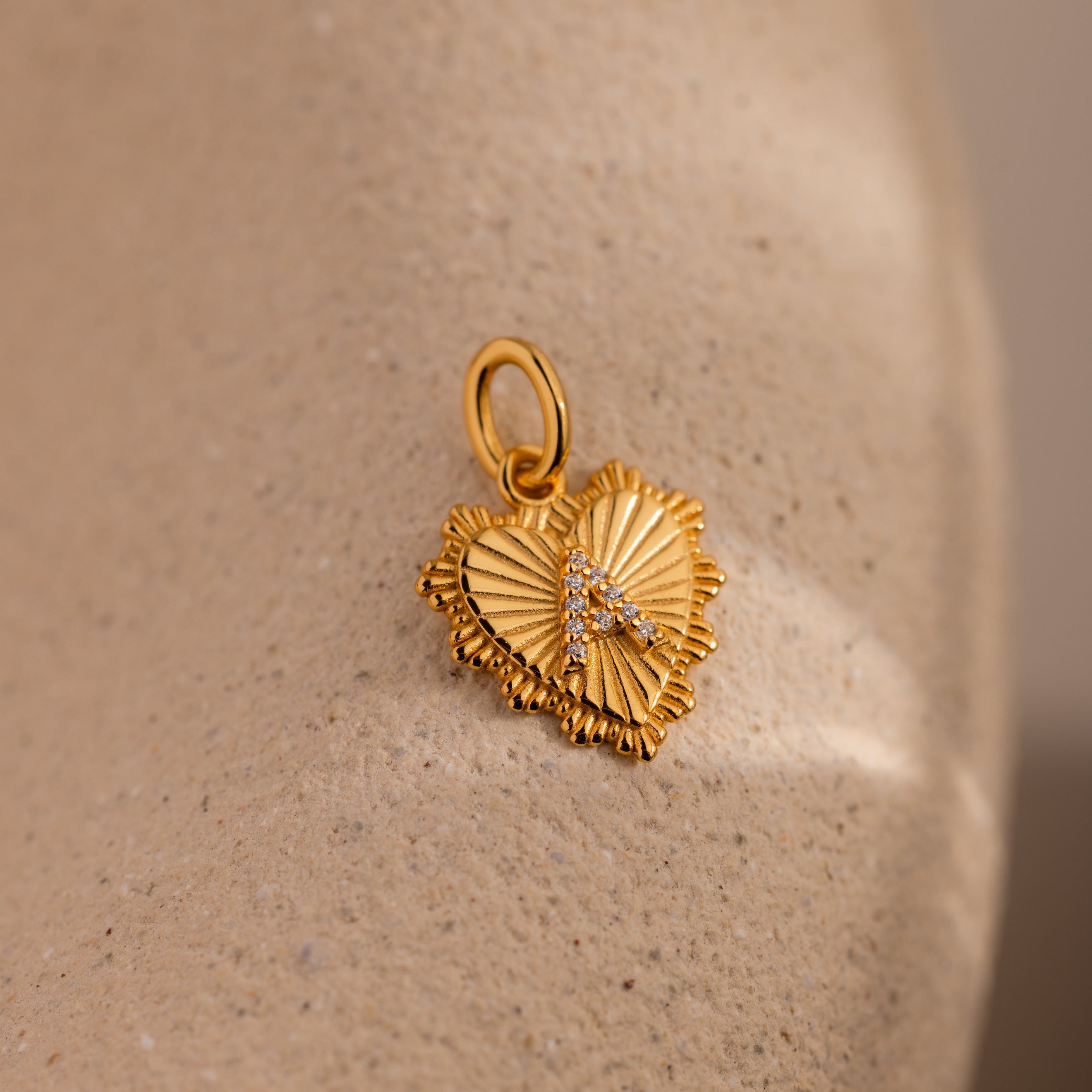 The Ribbed Heart Pave Initial Charm—a gold, heart-shaped pendant with textured ribbing and sparkling crystals—rests on a beige stone surface, making it an ideal choice for lovers of personalized jewelry.