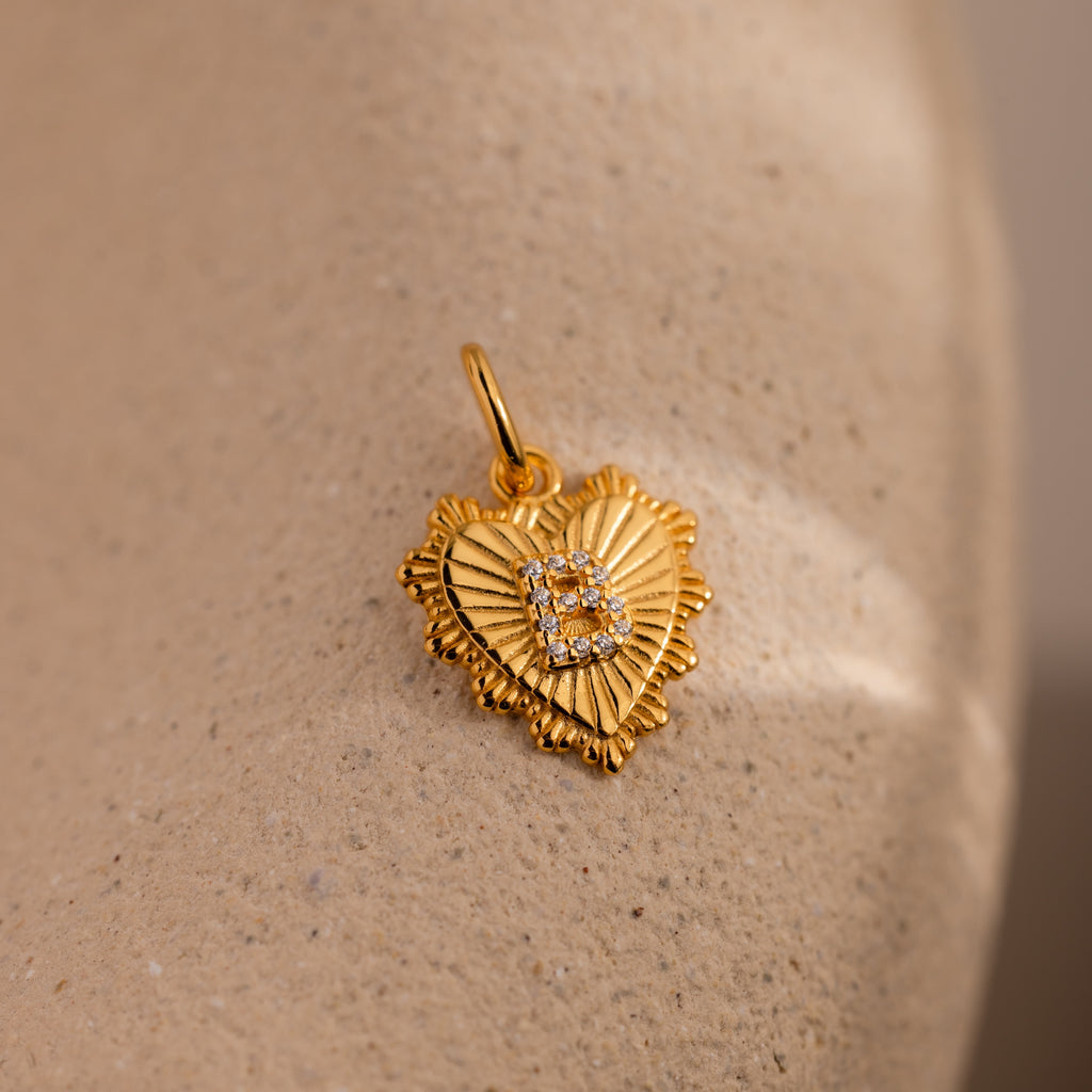 The Ribbed Heart Pave Initial Charm is a gold, heart-shaped pendant with textured edges and small diamonds, displayed on a beige stone—perfect for anyone who loves personalized jewelry.