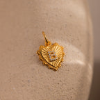 The Ribbed Heart Pave Initial Charm is a gold, heart-shaped pendant with textured edges and small diamonds, displayed on a beige stone—perfect for anyone who loves personalized jewelry.