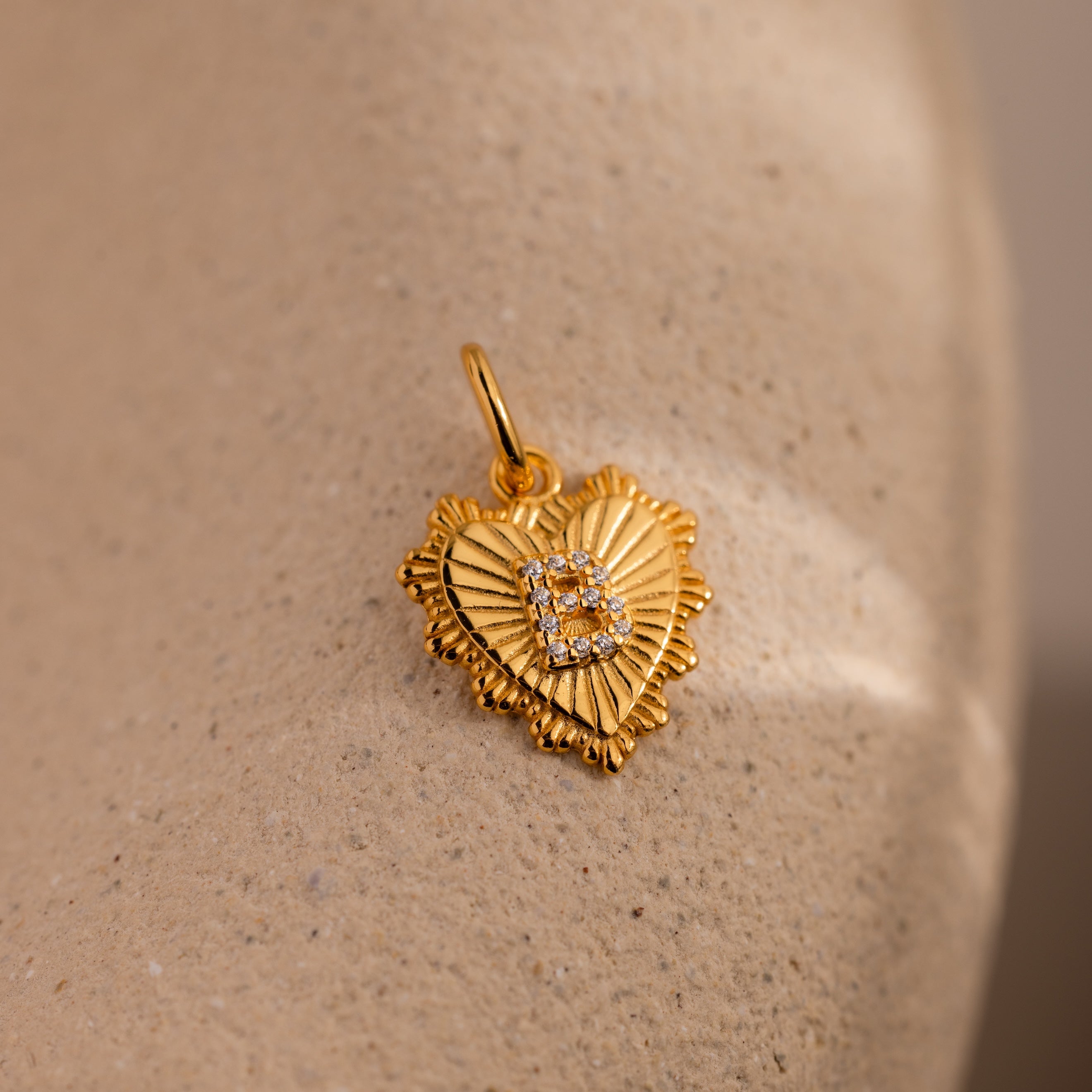 The Ribbed Heart Pave Initial Charm is a gold, heart-shaped pendant with textured edges and small diamonds, displayed on a beige stone—perfect for anyone who loves personalized jewelry.