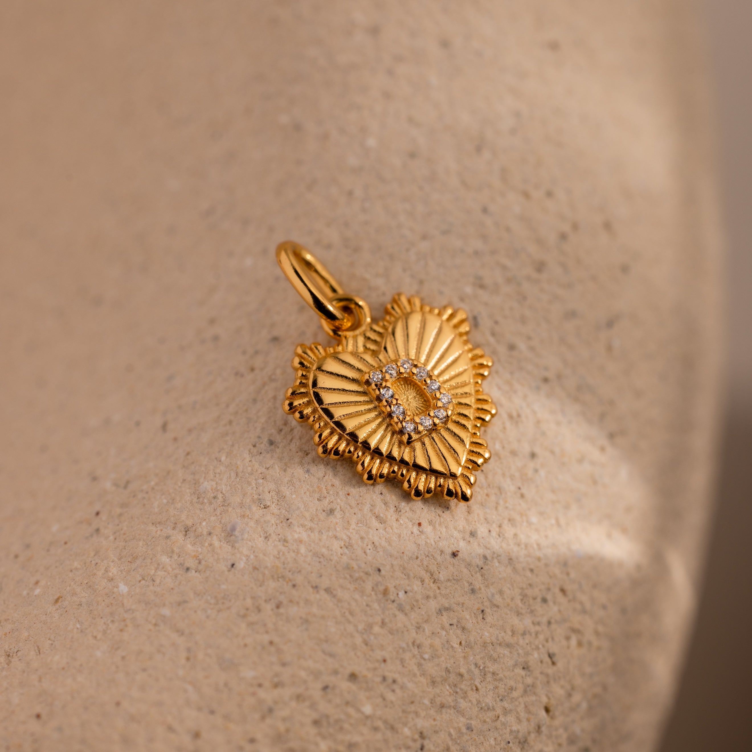 The Ribbed Heart Pave Initial Charm features a gold heart with a ribbed texture and a diamond-encrusted letter D, perfect for personalized jewelry. Shown on a beige surface.
