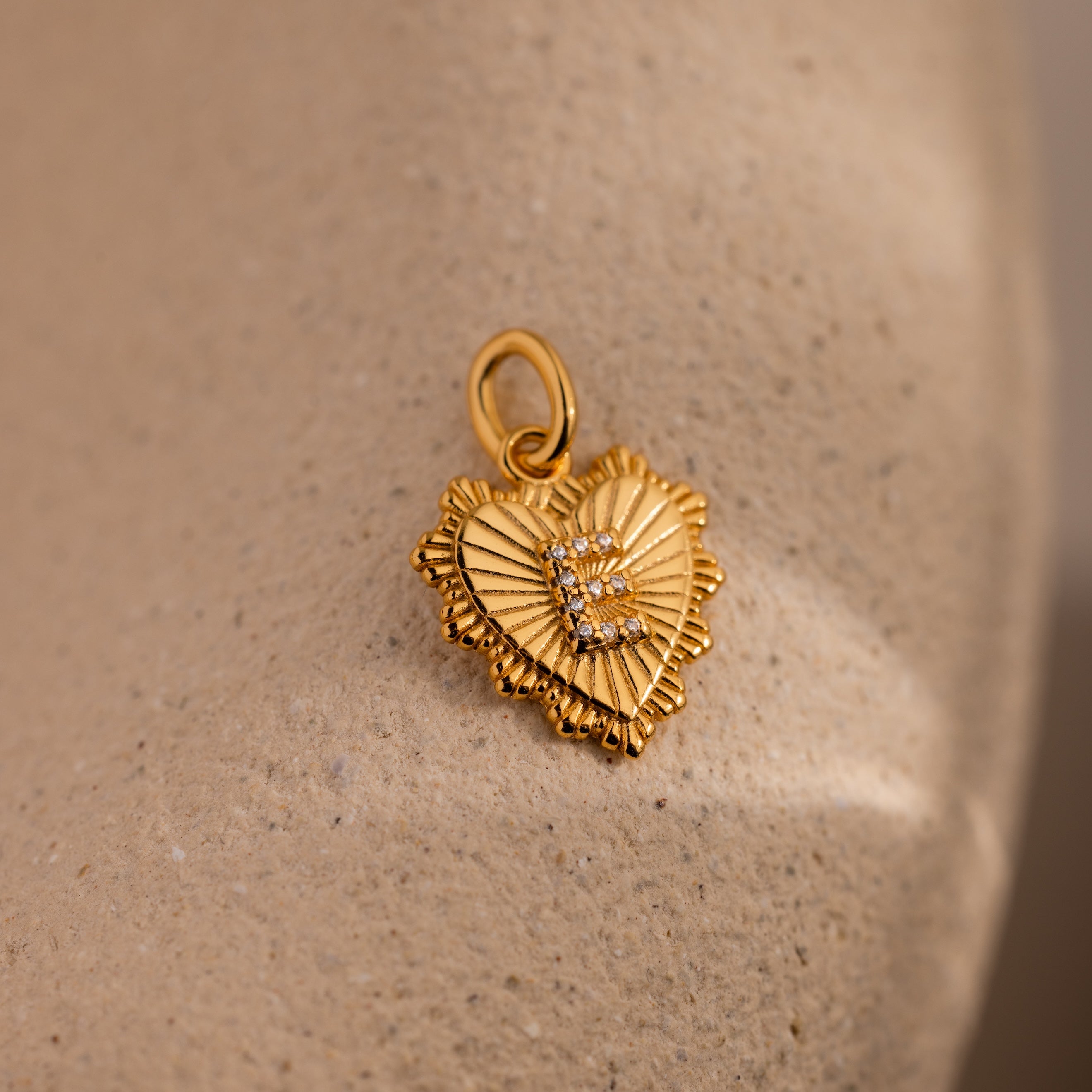The Ribbed Heart Pave Initial Charm features a gold heart with textured rays and clear stones, shown on a beige surface—ideal for anyone who loves personalized jewelry.