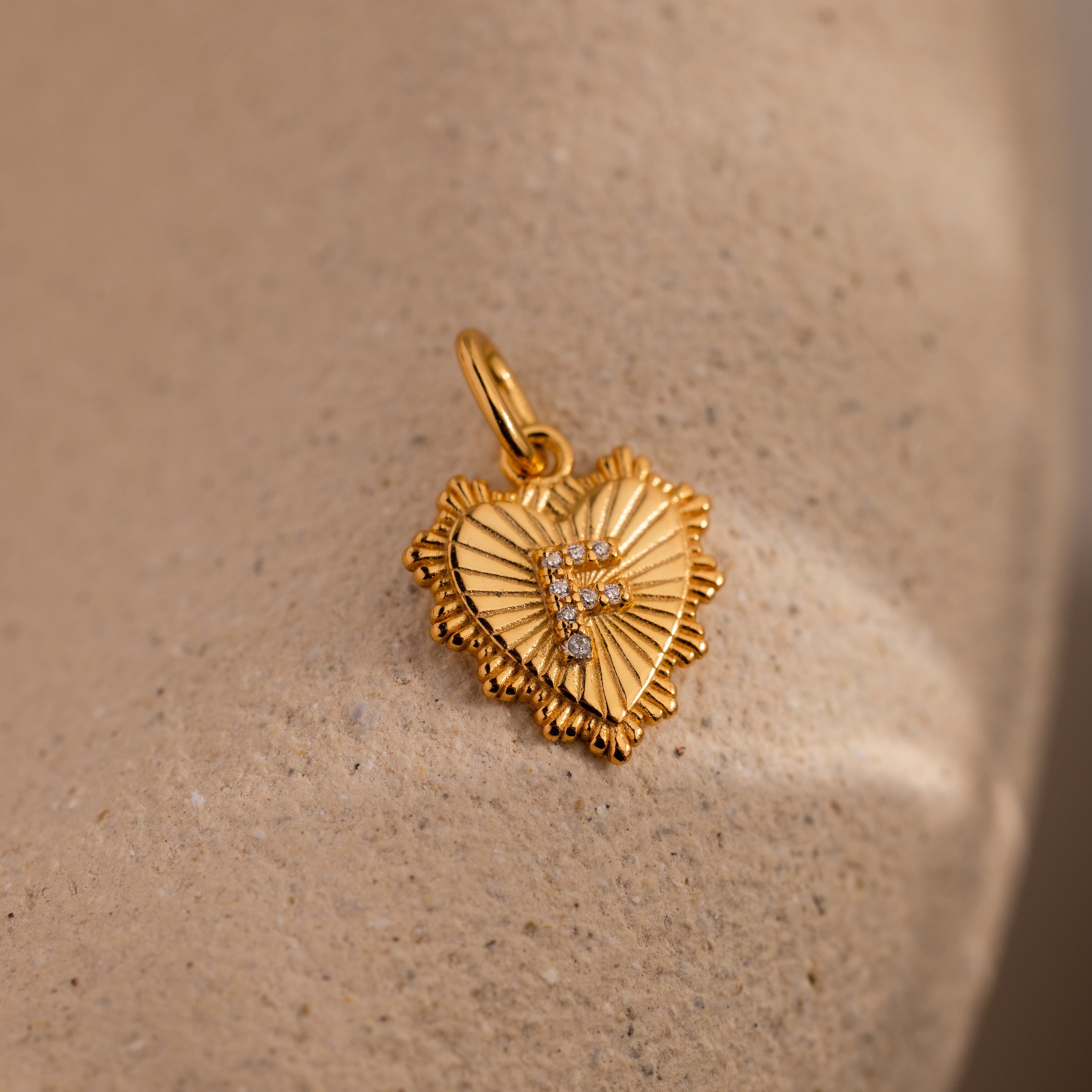 The Ribbed Heart Pave Initial Charm features diamond accents and rests on a textured beige surface, making it an elegant way to personalize your jewelry collection.