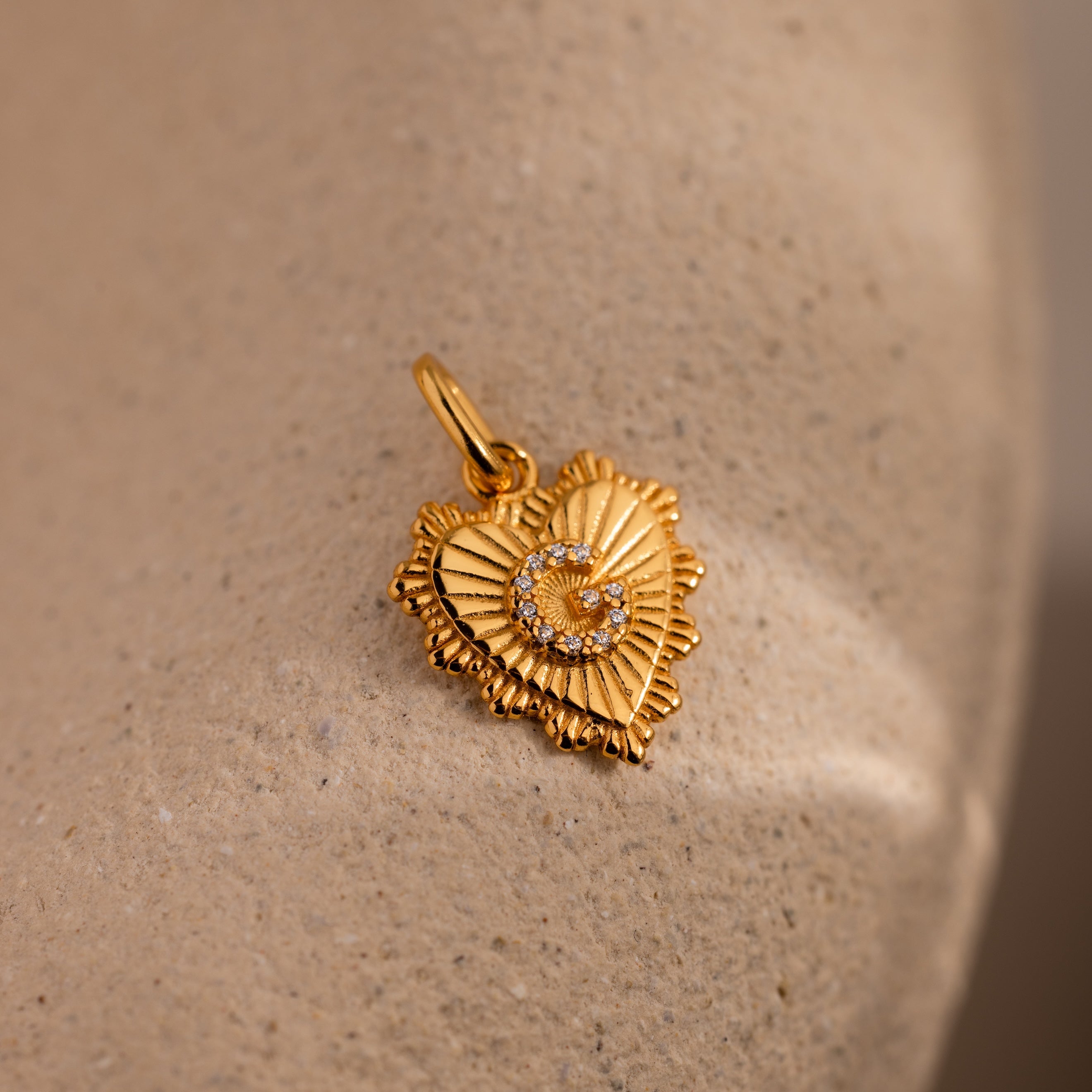 The Ribbed Heart Pave Initial Charm features a gold heart shape with textured edges and sparkling diamonds, making it a striking personalized jewelry piece shown on a beige stone surface.