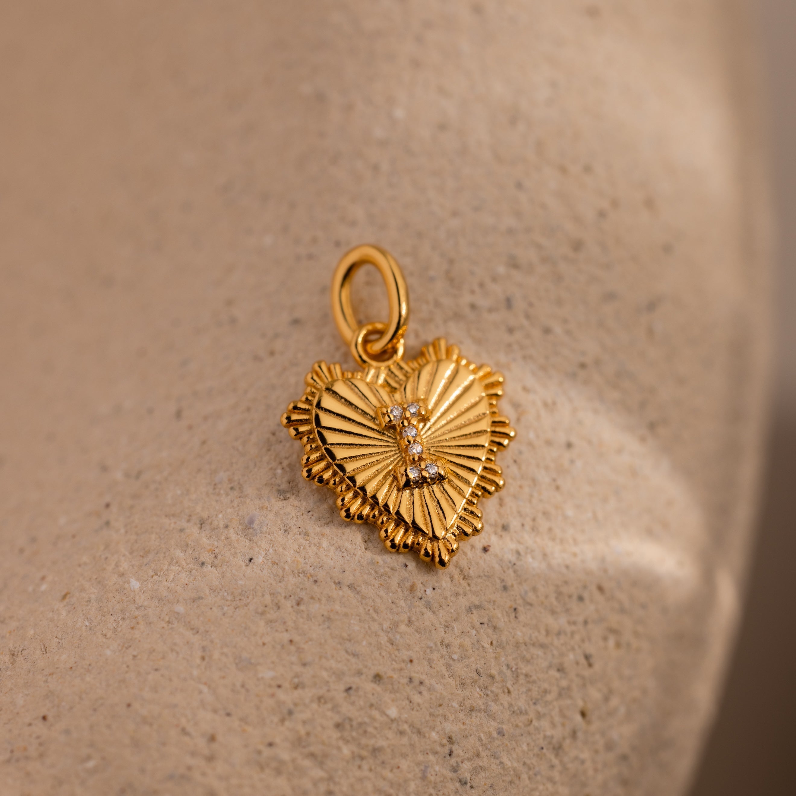 The Ribbed Heart Pave Initial Charm is a gold heart-shaped pendant with textured edges and central stones, shown on a beige textured surface—an elegant choice for those seeking personalized jewelry.