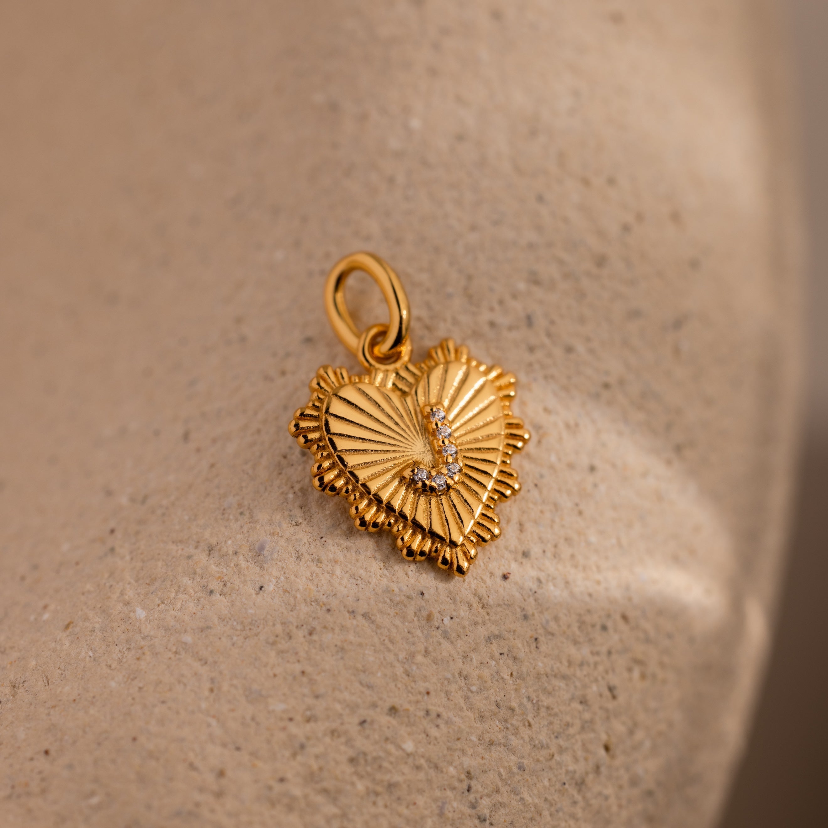 The Ribbed Heart Pave Initial Charm features a gold heart shape with textured edges and sparkling pave details, offering an elegant personalized jewelry option beautifully shown on a beige surface.