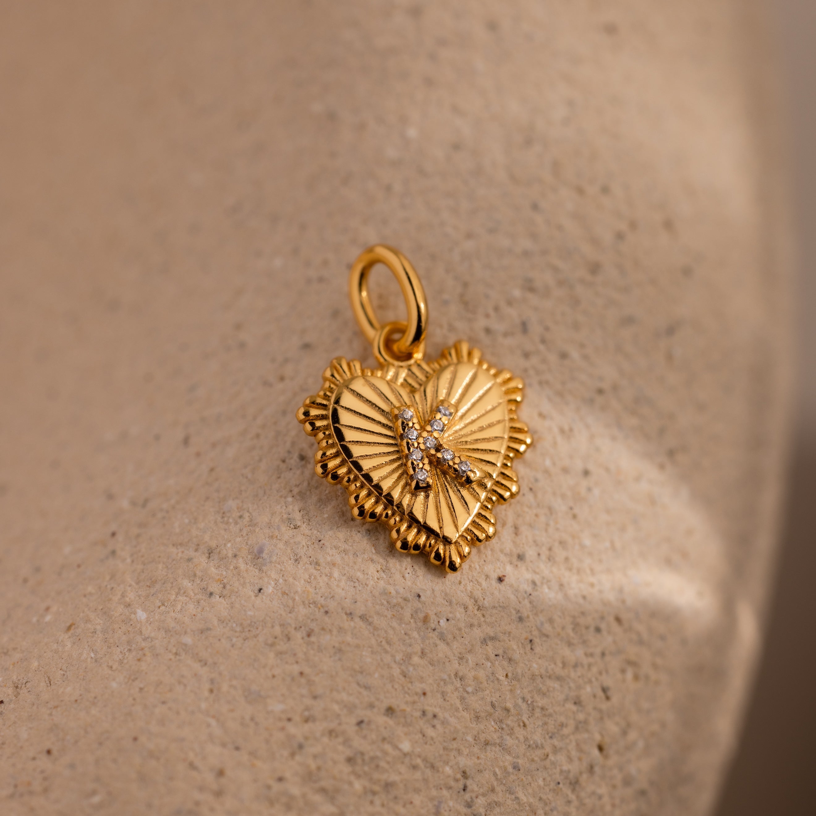 The Ribbed Heart Pave Initial Charm features a gold heart design with textured edges and sparkling accents, reminiscent of a personalized jewelry piece, beautifully showcased on a sandy surface.