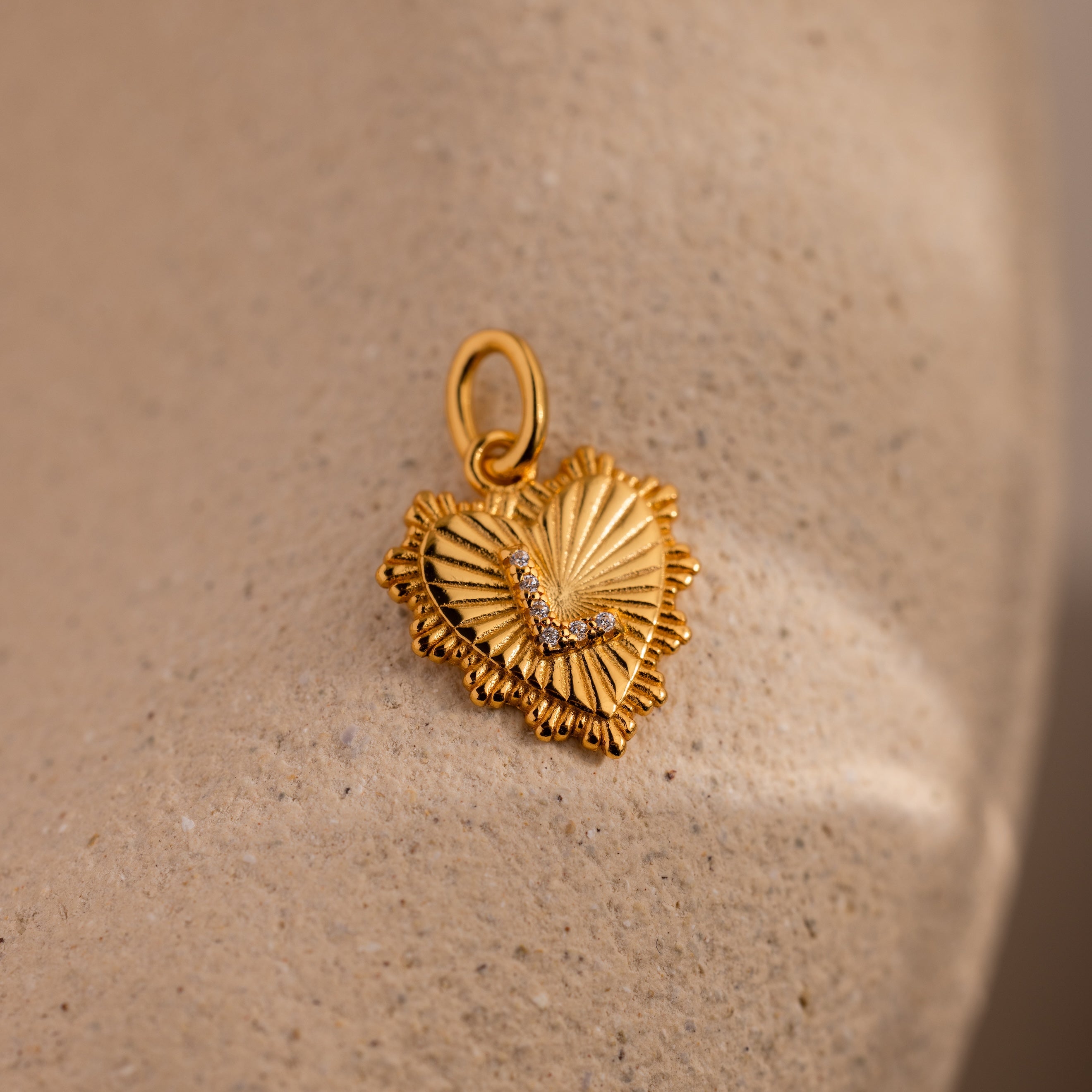 The Ribbed Heart Pave Initial Charm features textured details and sparkling gems, displayed on a beige textured surface—an ideal choice for fans of personalized jewelry.