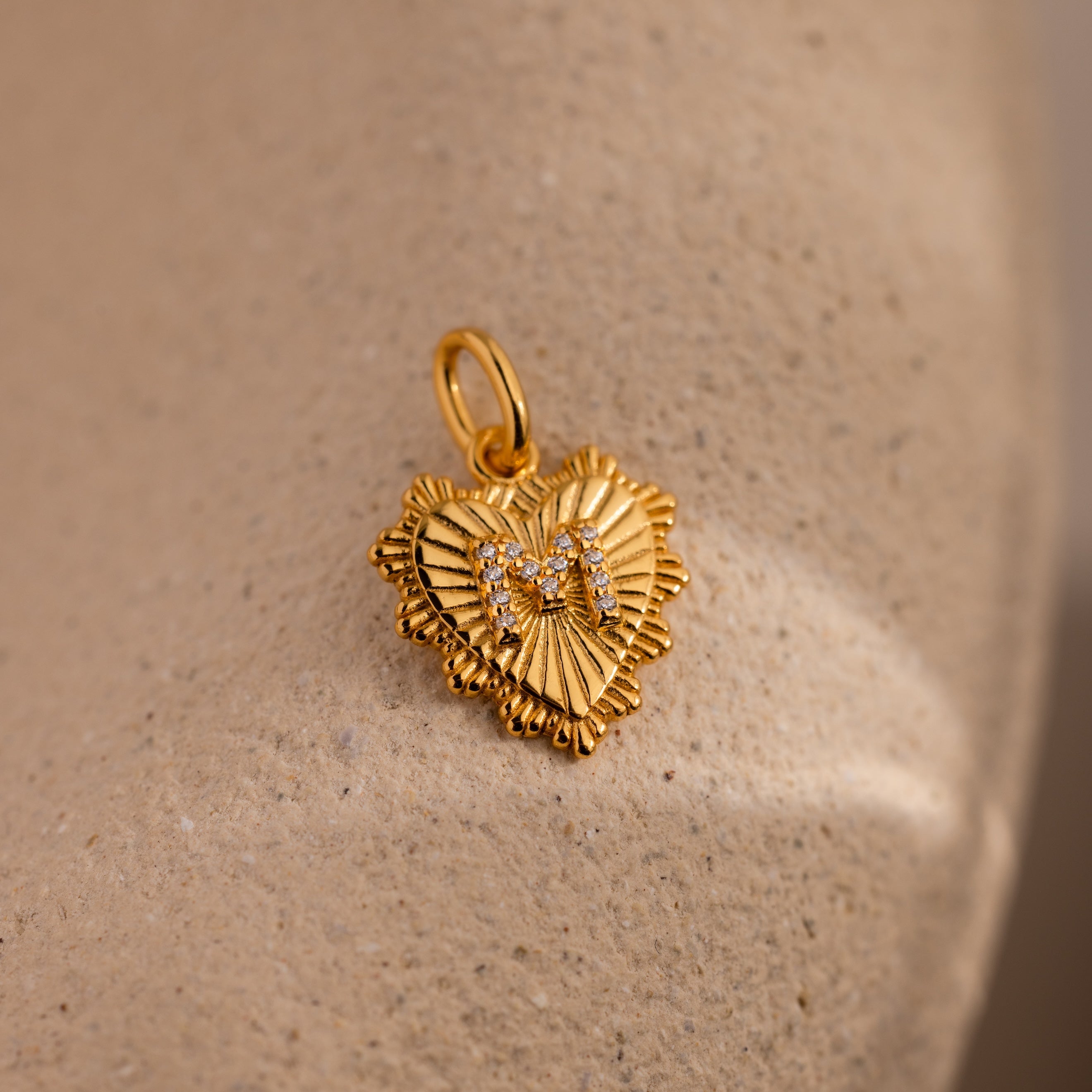 The Ribbed Heart Pave Initial Charm features a gold heart shape with ribbed edges and sparkling stones, offering personalized elegance—perfectly showcased on a beige surface.