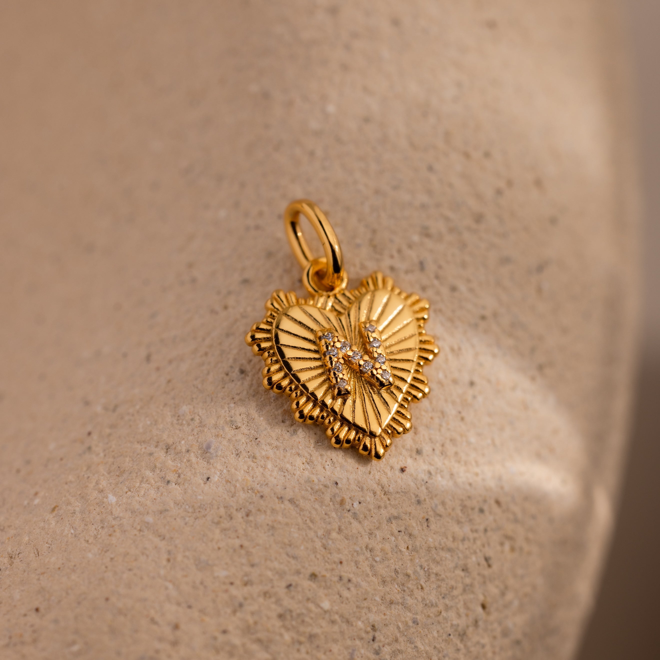 The Ribbed Heart Pave Initial Charm features a gold heart shape with ribbed edges and sparkling stones, making it a stunning personalized piece, showcased on a sandy beige background.