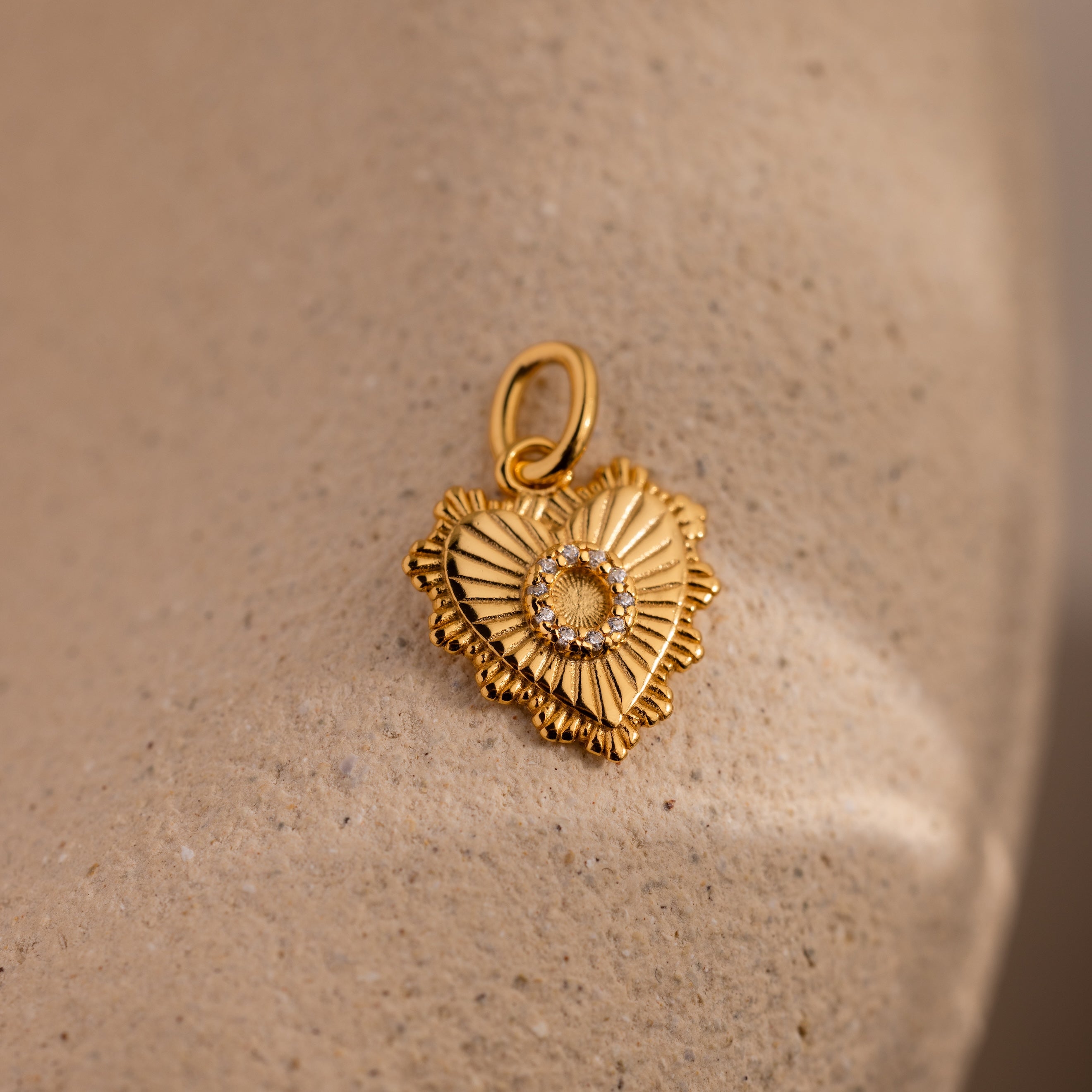 The Ribbed Heart Pave Initial Charm features a gold heart-shaped design with a central gem and textured details, displayed on a beige stone surface—ideal for fans of personalized jewelry.