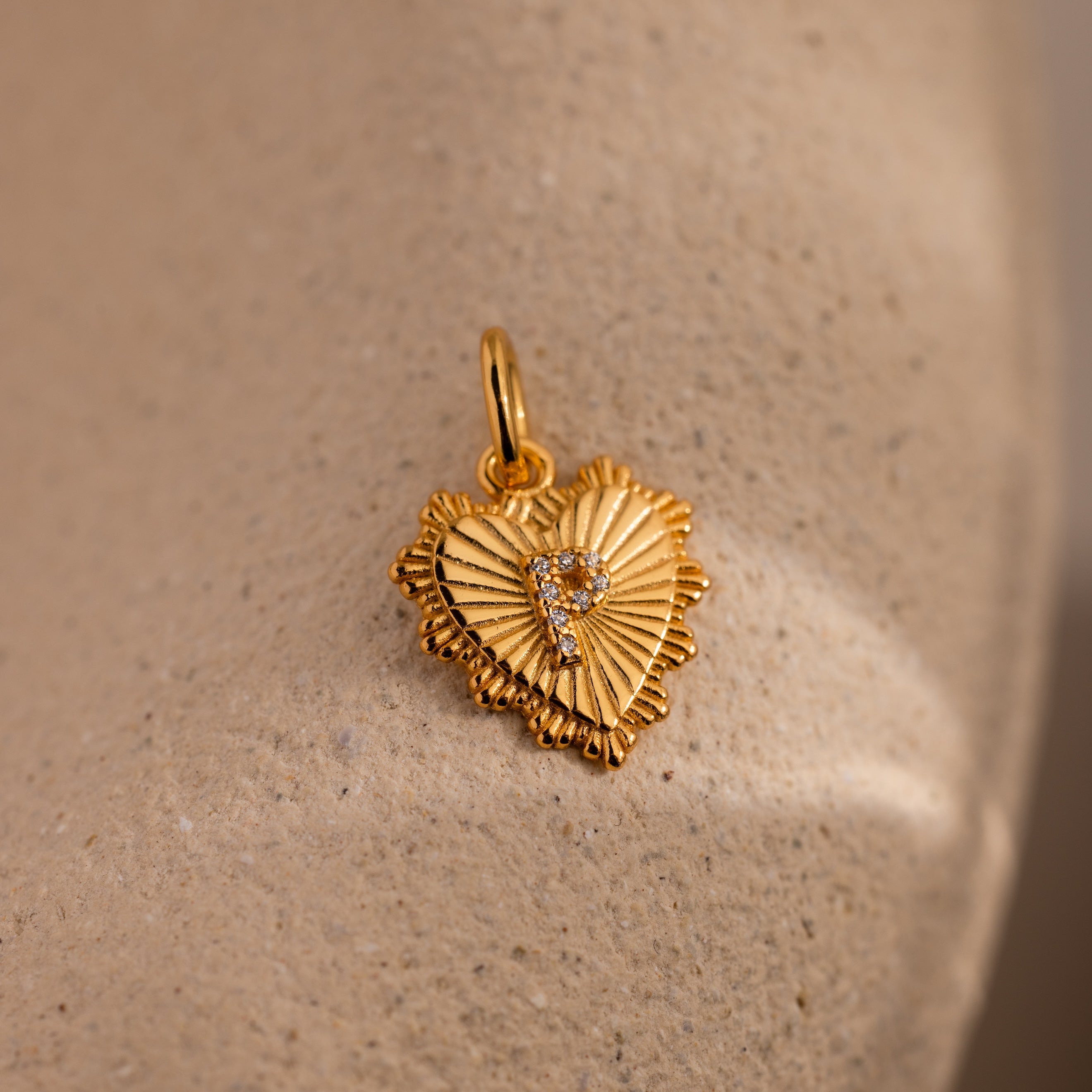 The Ribbed Heart Pave Initial Charm features a gold heart design with textured edges and a gem-studded center, offering an elegant, personalized addition to any jewelry collection.