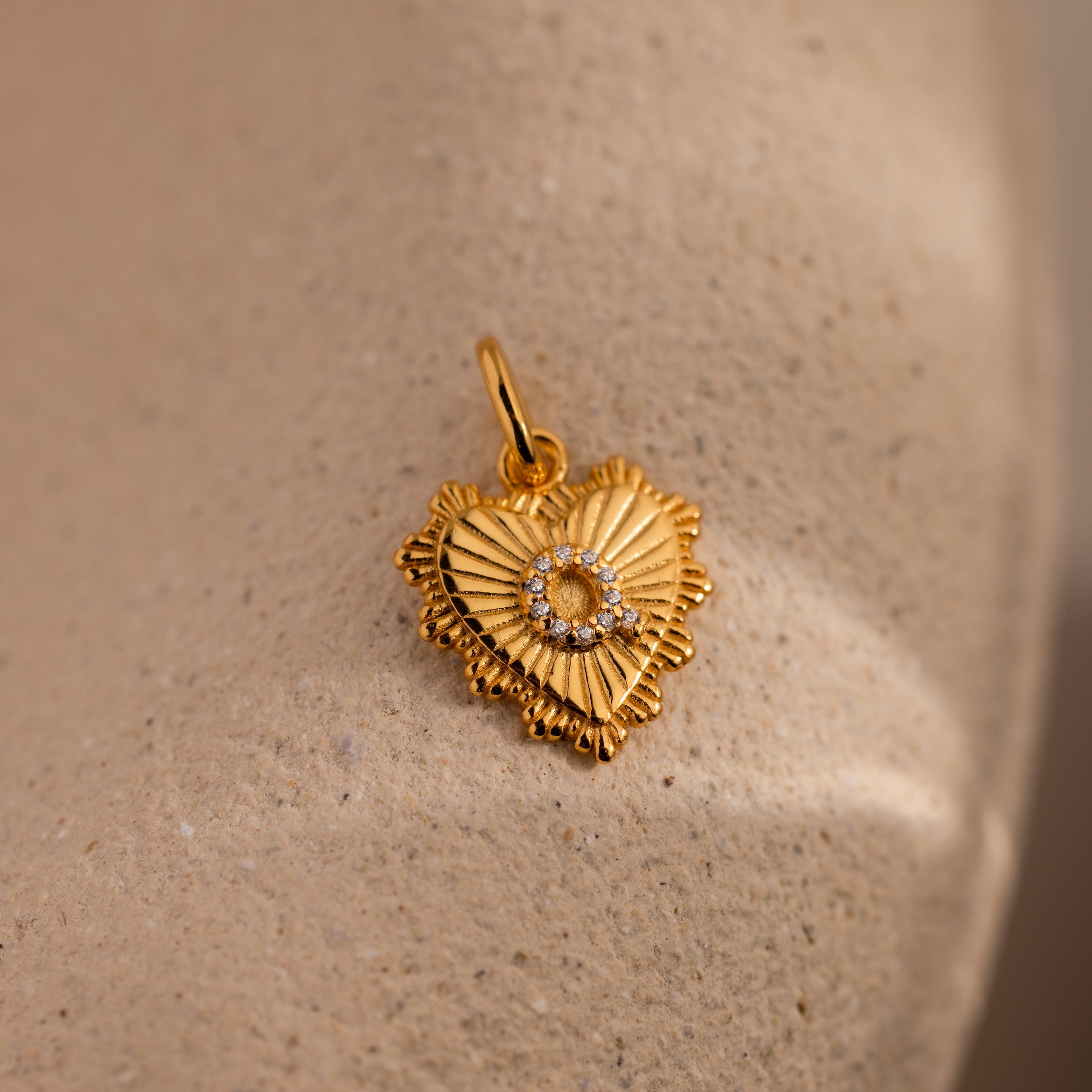 The Ribbed Heart Pave Initial Charm showcases textured rays and a central ring of sparkling stones, elegantly displayed on a beige surface—an ideal choice for lovers of personalized jewelry.