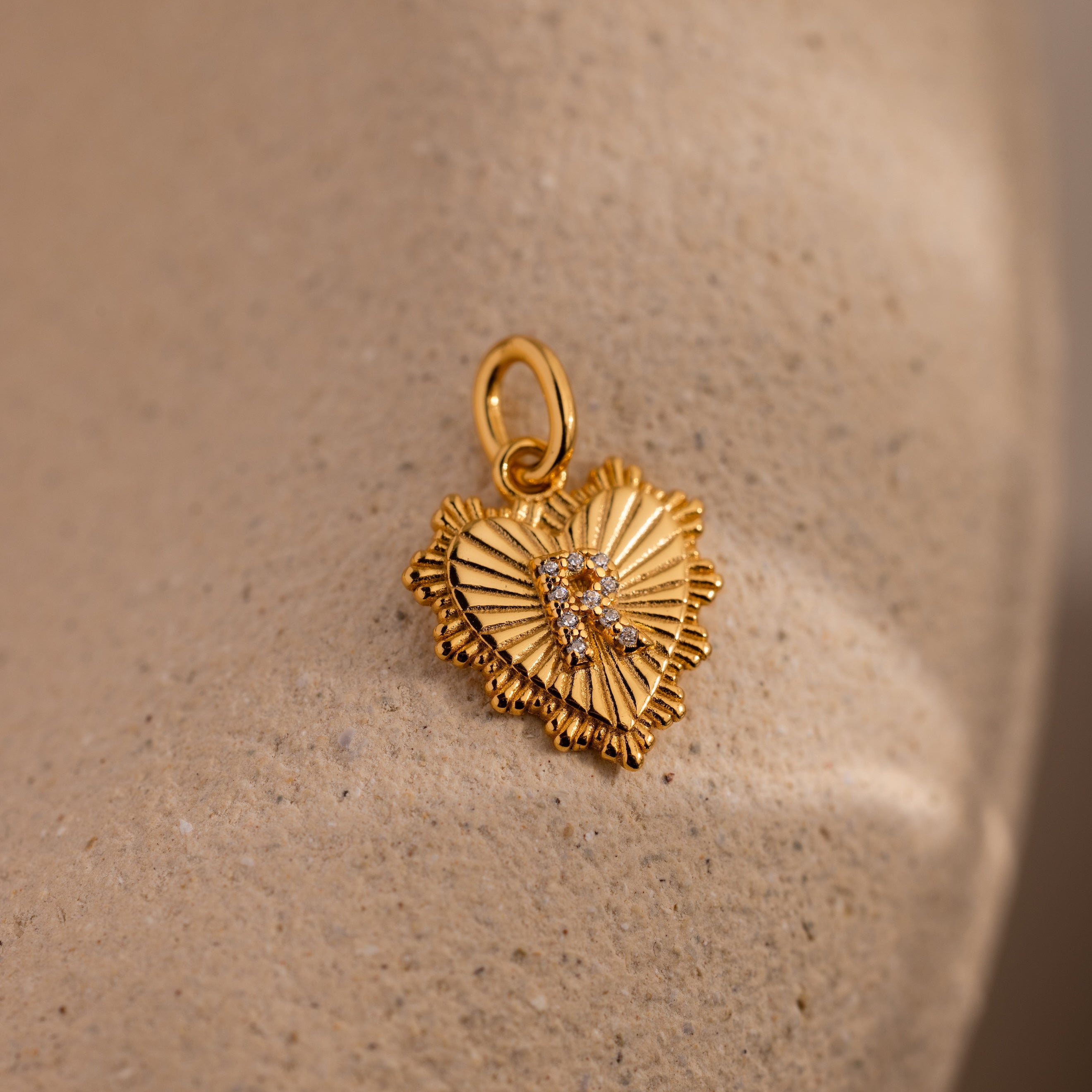 The Ribbed Heart Pave Initial Charm features a gold heart-shaped pendant with textured rays and sparkling diamonds, offering elegant personalized jewelry, shown on a beige stone surface.