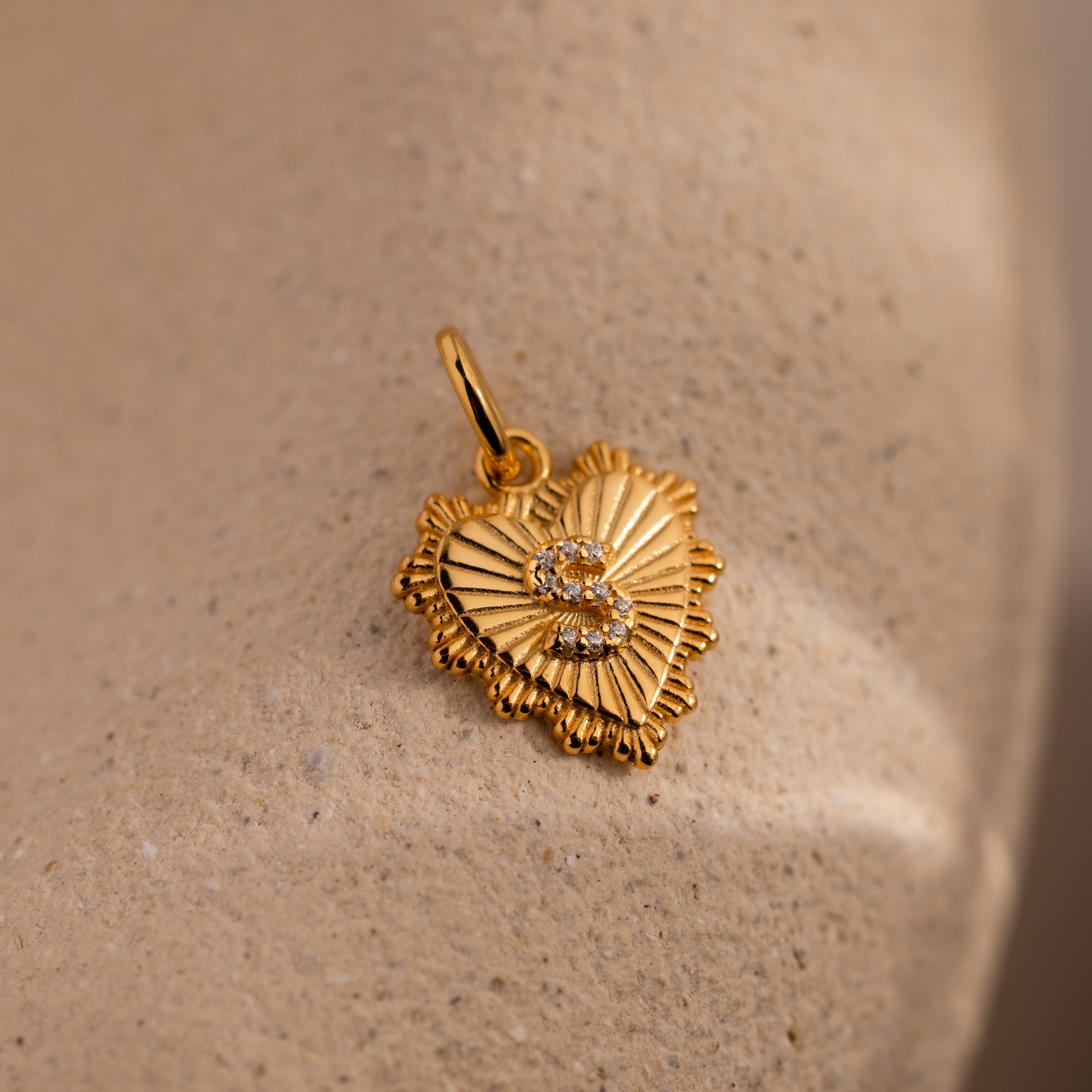 The Ribbed Heart Pave Initial Charm features a gold heart shape with ribbed rays and small sparkling jewels, displayed on a beige textured surface.