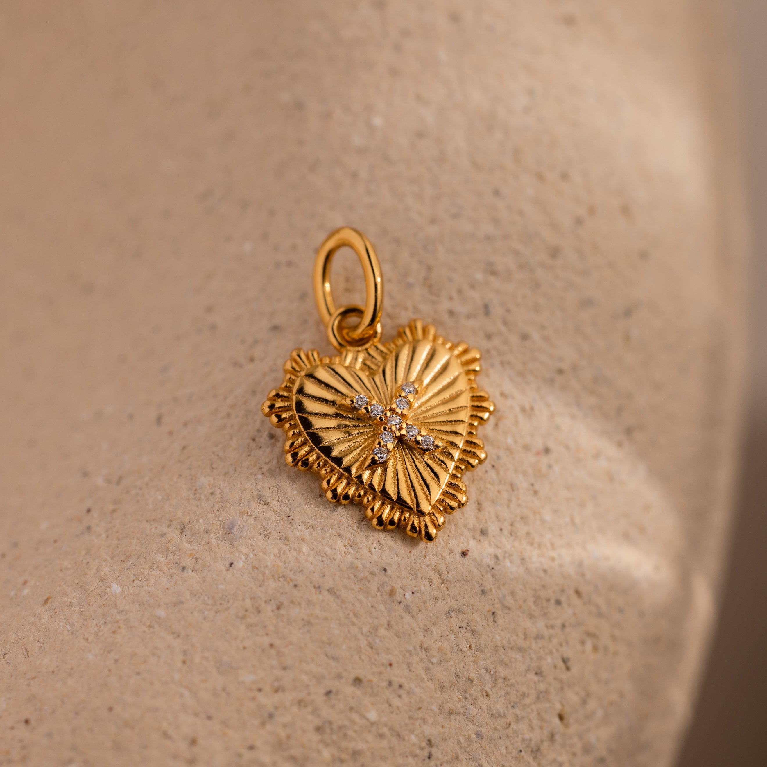 The Ribbed Heart Pave Initial Charm is a gold heart-shaped pendant with a textured surface and sparkling diamonds, perfect for lovers of personalized jewelry. It’s elegantly displayed on a beige textured background.