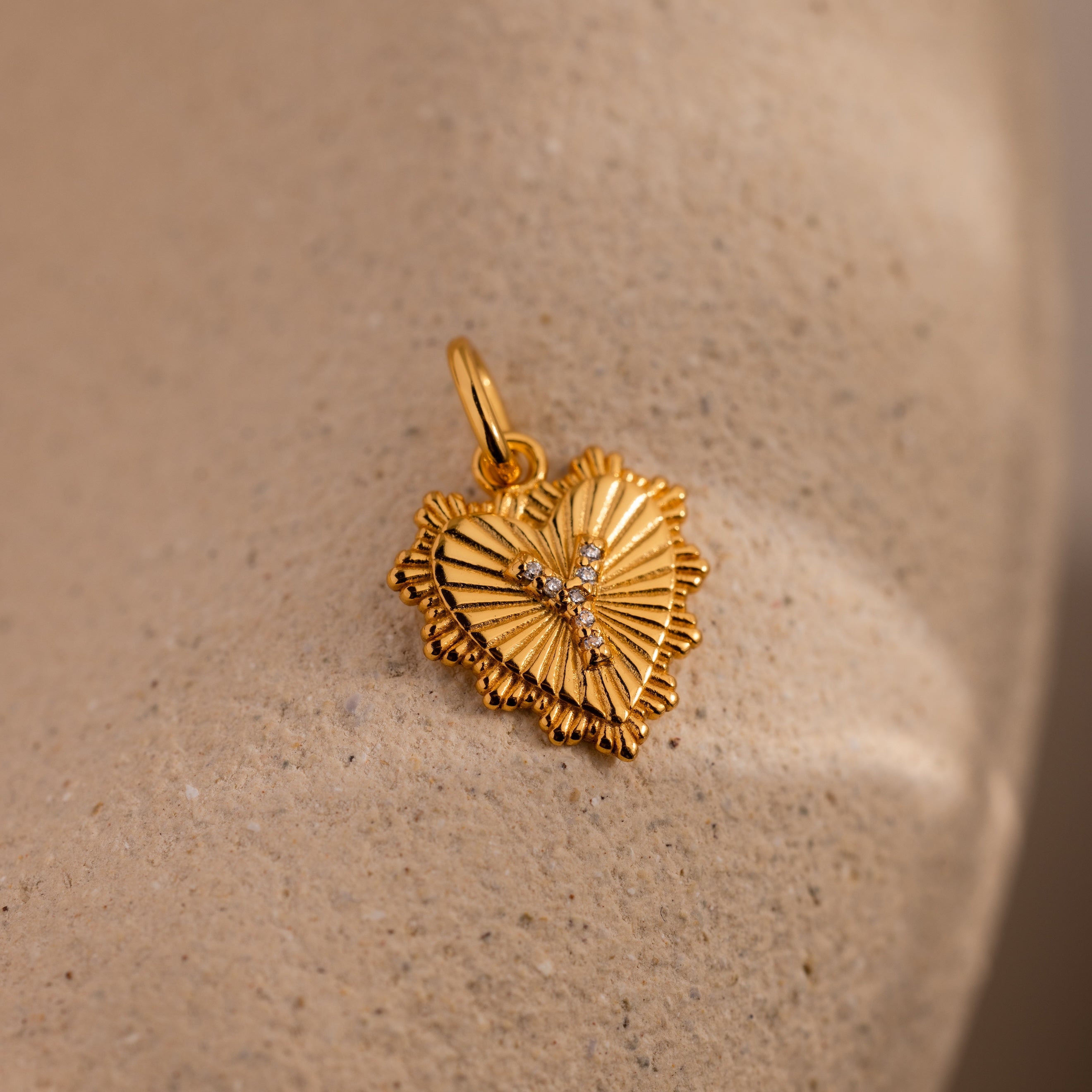The Ribbed Heart Pave Initial Charm features textured edges and sparkling gems, displayed on a beige stone surface—a perfect personalized jewelry piece.