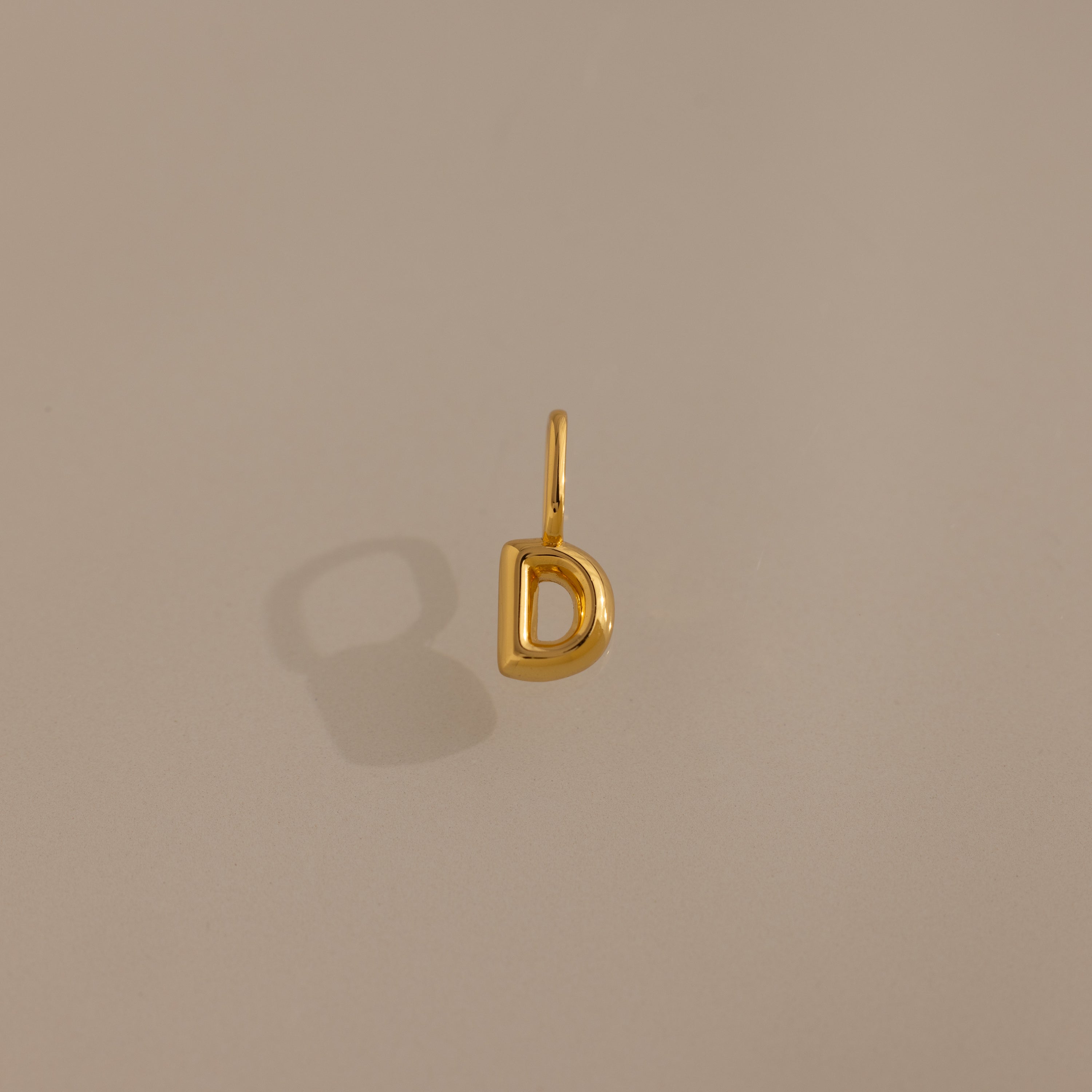 Bubble Letter Charm in 18K Gold D pendant displayed on a neutral beige background with a soft left shadow—ideal personalized jewelry or a charming addition for bracelet enthusiasts.