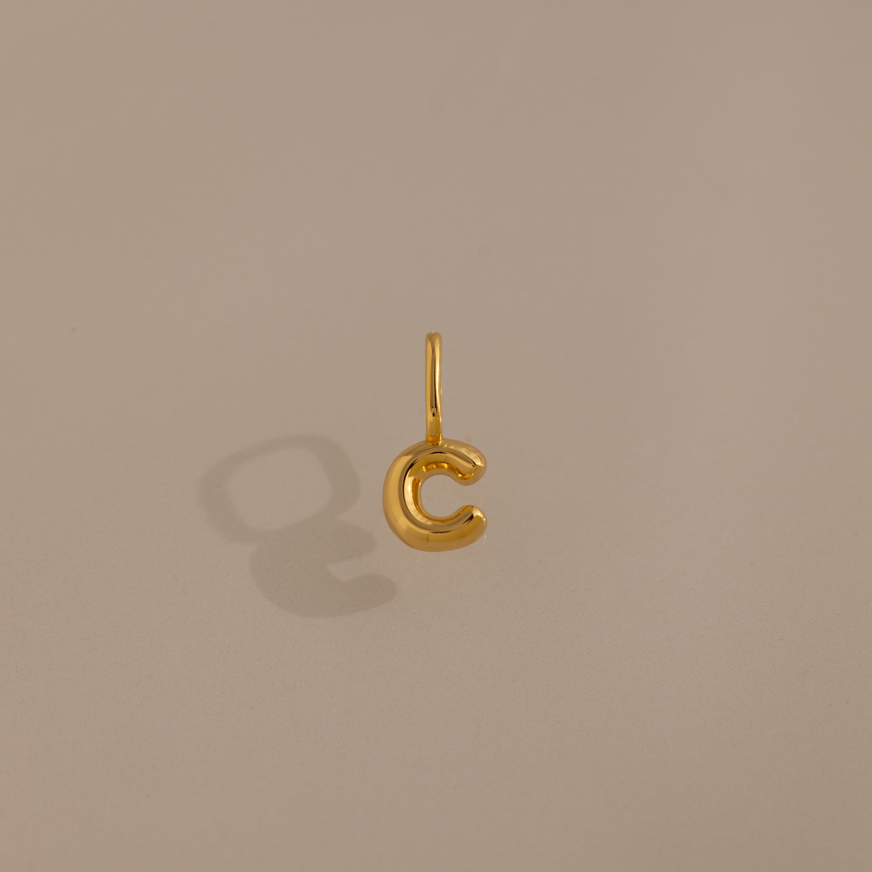 The Bubble Letter Charm in 18K Gold shaped like the letter C casts a shadow on a beige background, making it an ideal personalized jewelry piece or bracelet charm.