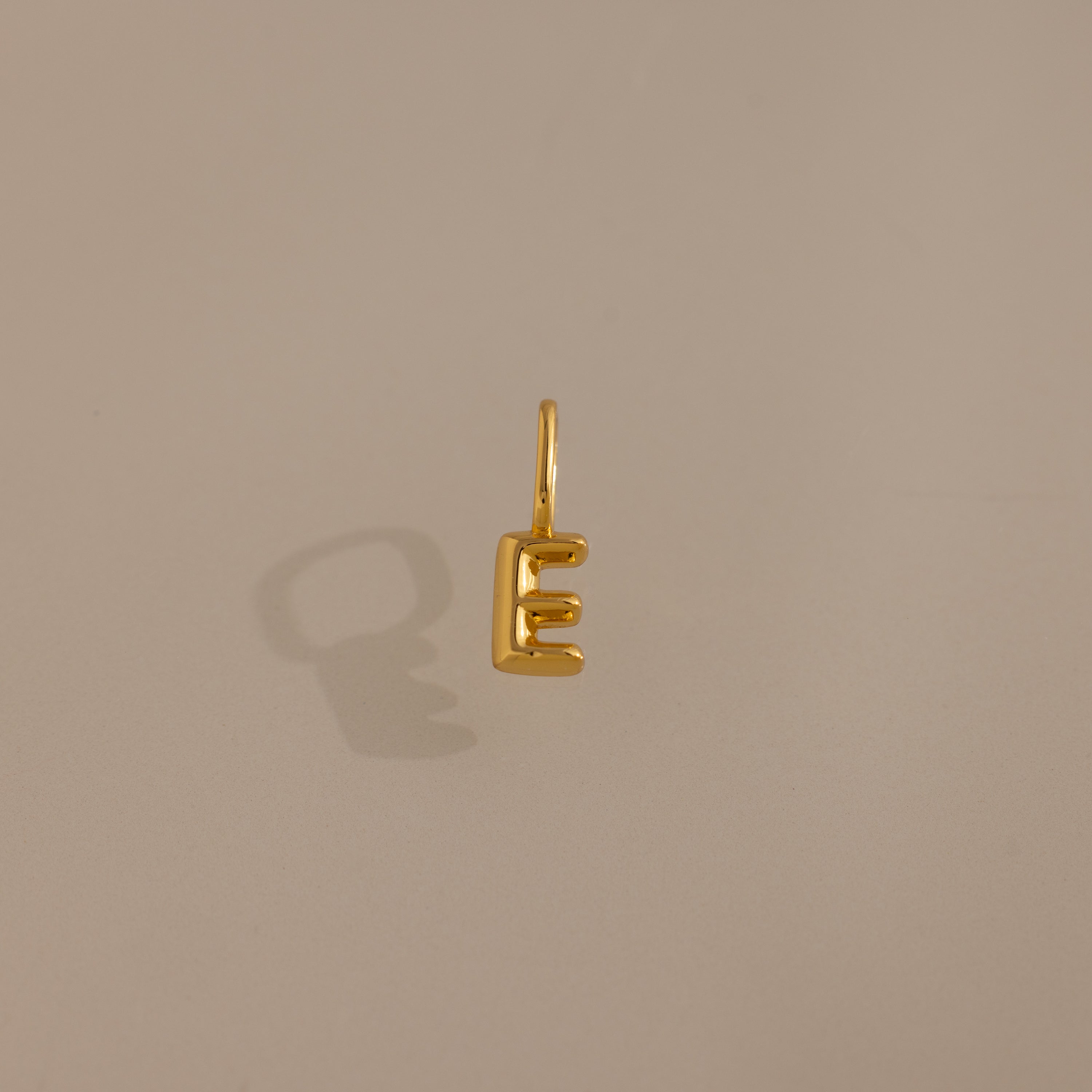 The Bubble Letter Charm in 18K Gold, shaped like the letter E, casts a shadow on a plain beige background—an elegant piece of personalized jewelry.