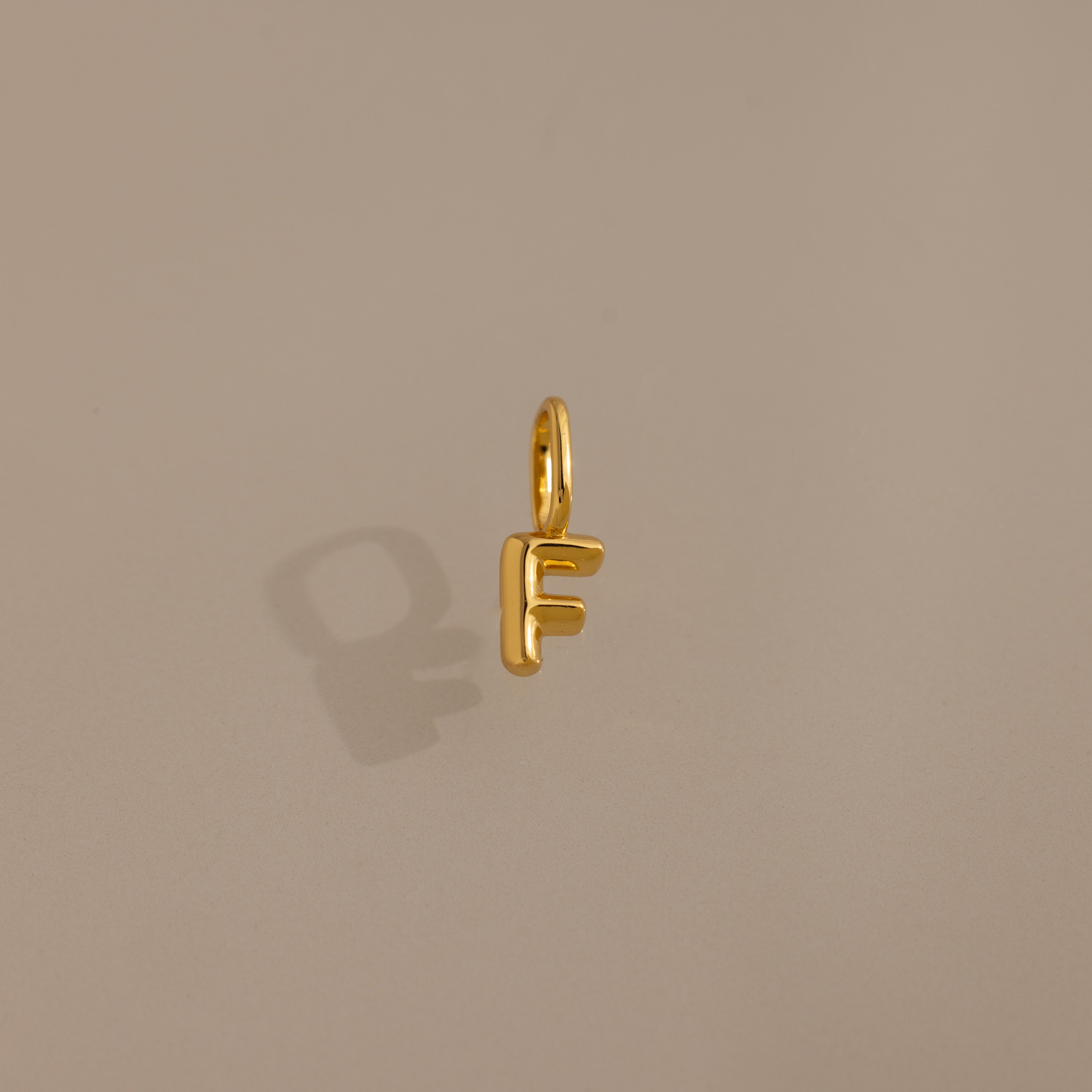 The Bubble Letter Charm in 18K Gold, shaped like the letter F, sits on a beige background—ideal as personalized jewelry or a distinctive charm for bracelets.